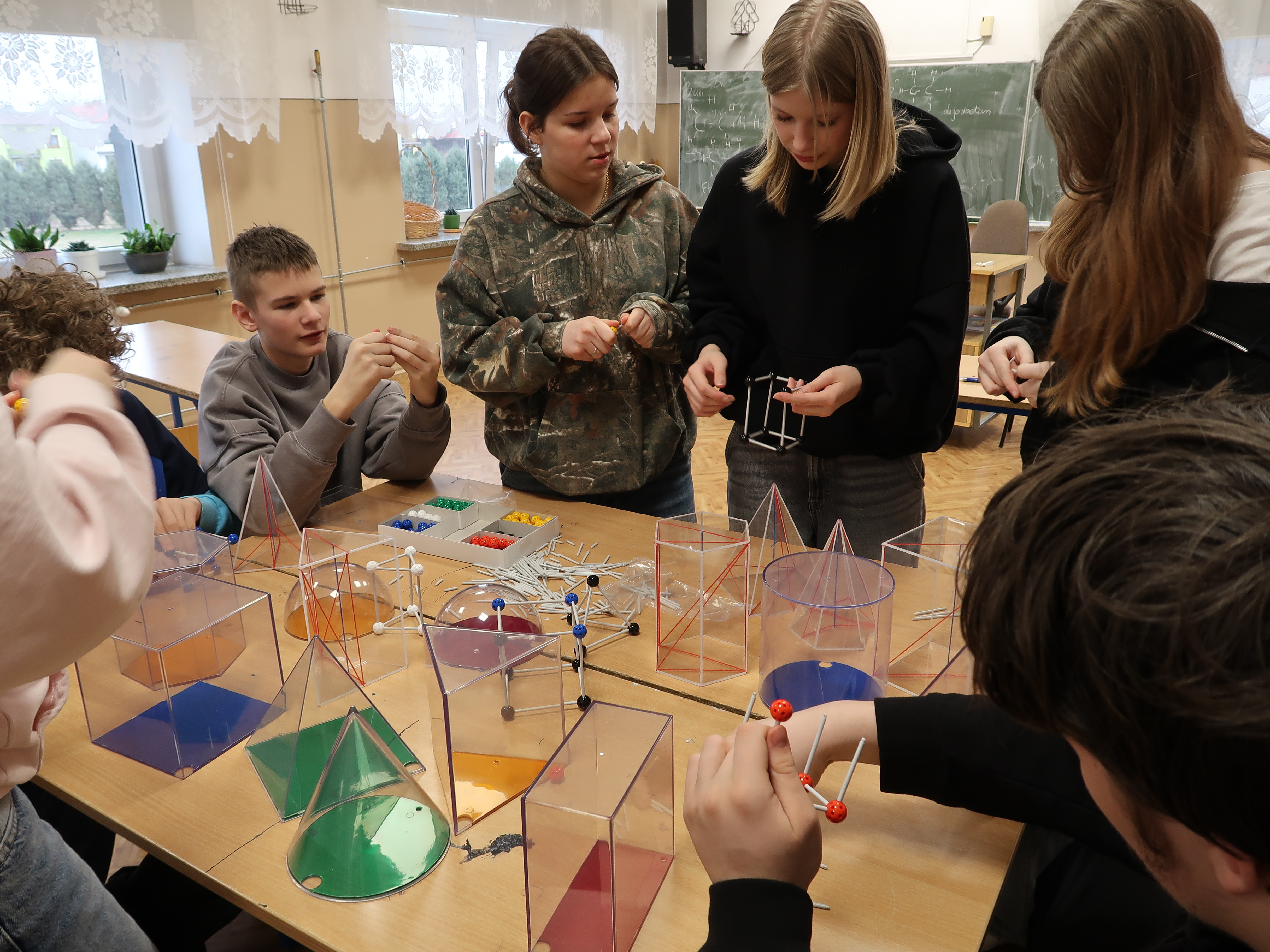Grupa nastolatków przy stole z przezroczystymi modelami brył geometrycznych w klasie z tablicą i oknami w tle.