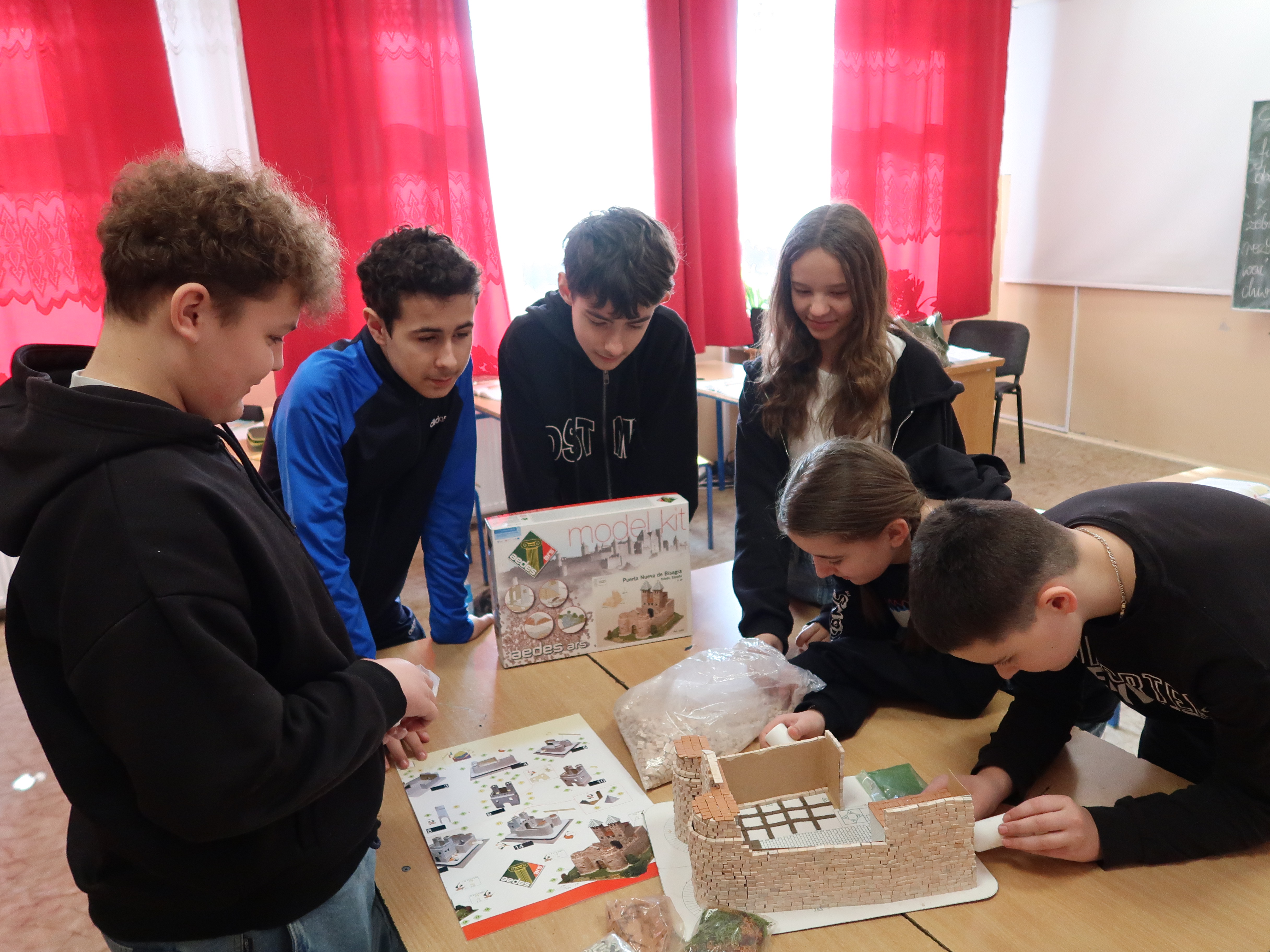Grupa młodzieży przy stole z modelem zamku, instrukcją i pudełkiem zestawu do składania, w tle czerwone zasłony i jasne ściany.