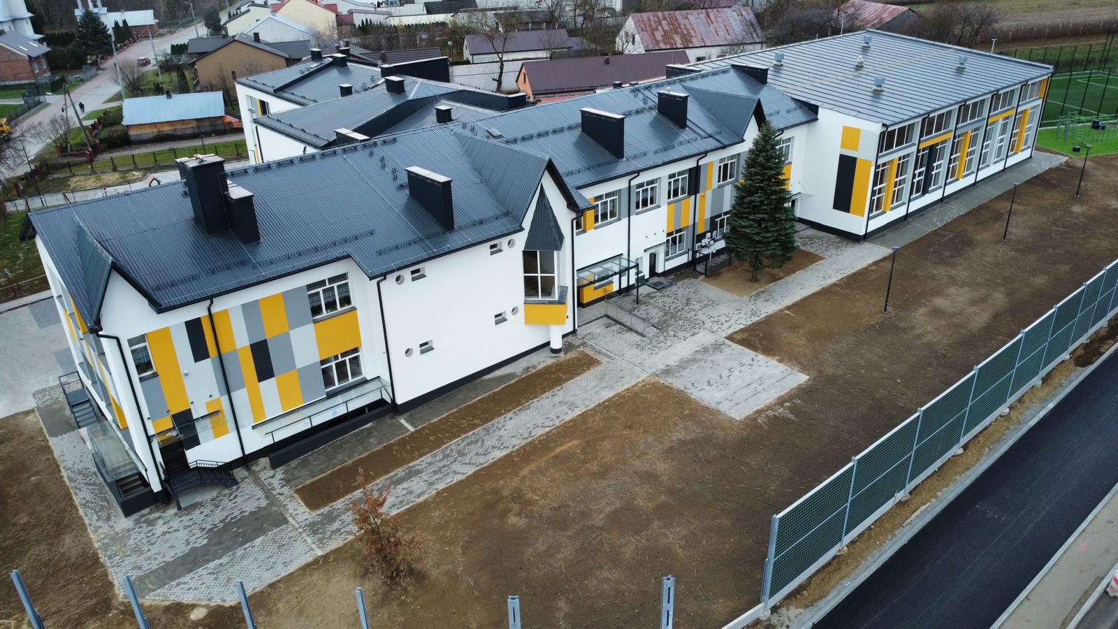 Biały budynek z płaskim i spadzistym dachem, elewacja z żółtymi, szarymi i czarnymi prostokątami, chodnik z szarych kostek, ogrodzenie metalowe, drzewa i trawa.