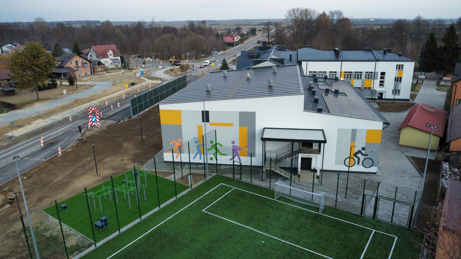 Budynek sportowy z muralem kolorowych sylwetek uprawiających różne dyscypliny, zielone boisko piłkarskie z białymi liniami i ogrodzeniem, otoczenie drzew i domów.