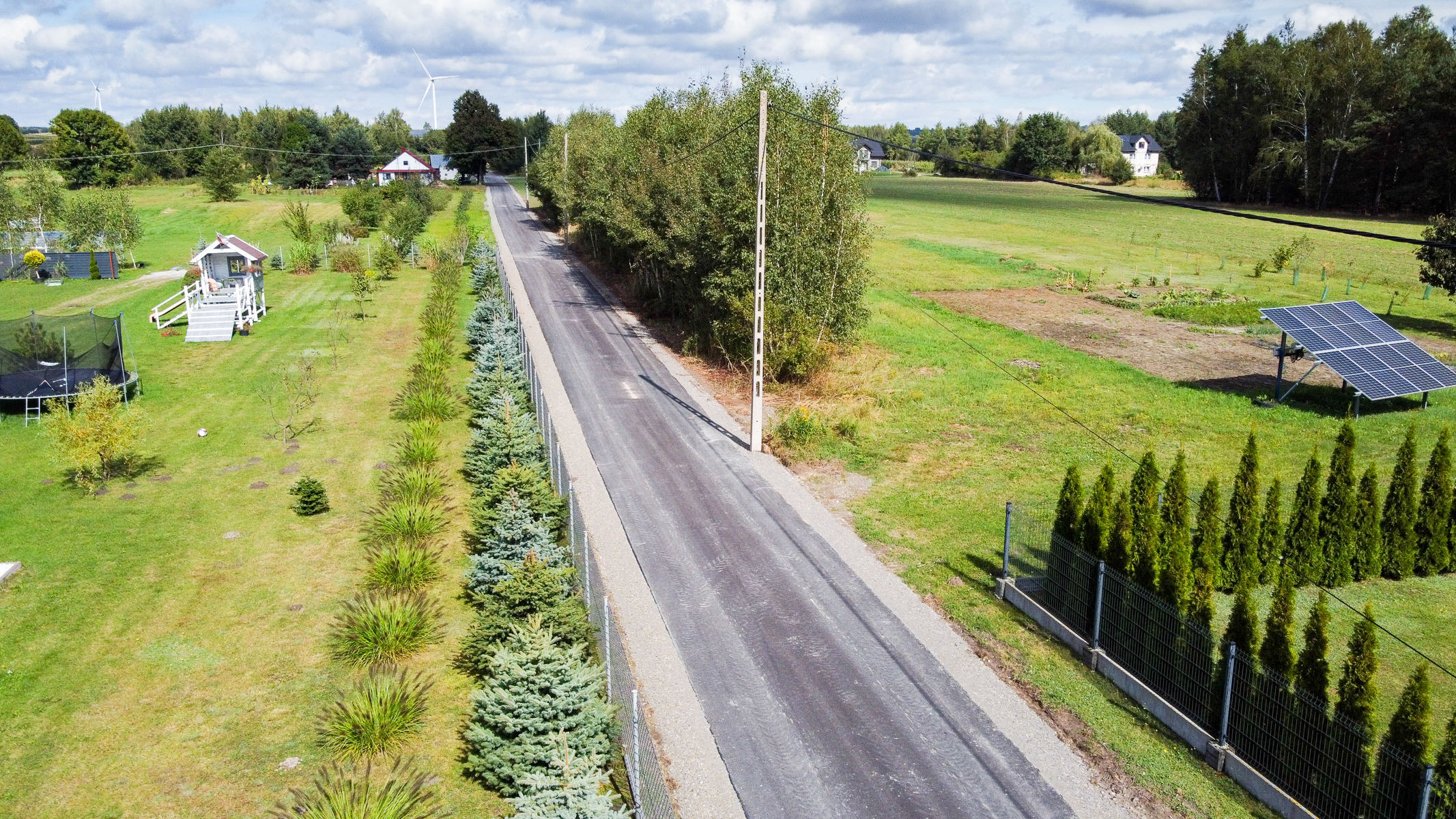 Wąska asfaltowa droga między dwoma ogrodzonymi działkami z zieloną trawą, drzewami, krzewami, domkiem na palach, trampoliną i panelami słonecznymi pod niebieskim niebem.