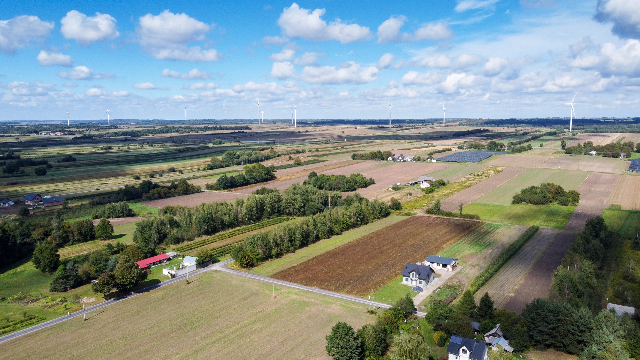 Widok z lotu ptaka na pola uprawne o różnych kolorach, kilka domów, drogi, skupiska drzew oraz wiatraki na horyzoncie pod niebieskim niebem z chmurami.