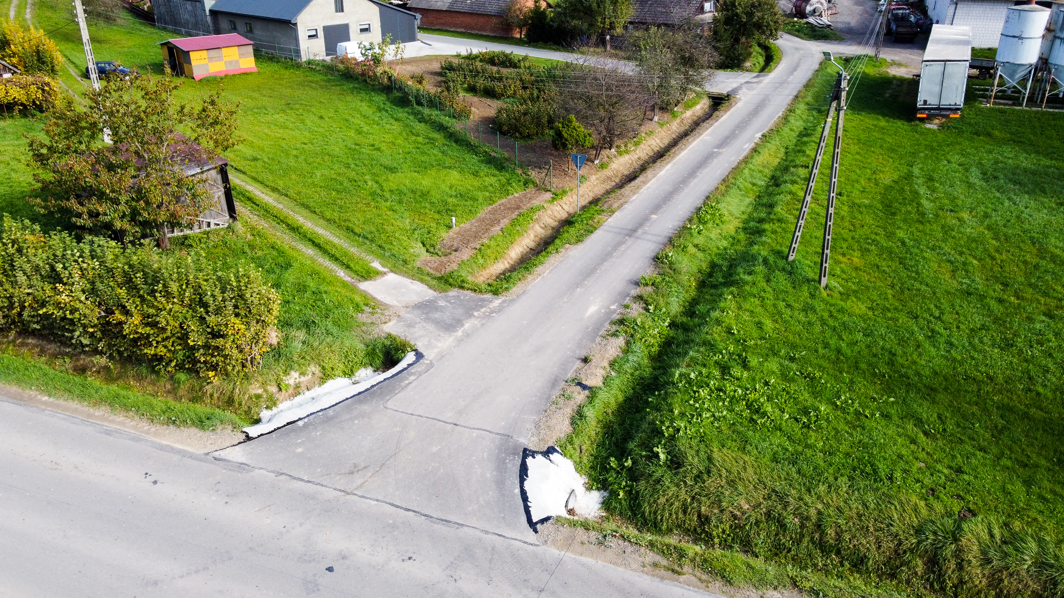 Rozgałęzienie asfaltowej drogi na tle zielonych trawników, przydrożne słupy energetyczne, zabudowania gospodarcze i ogrodzony ogródek warzywny.