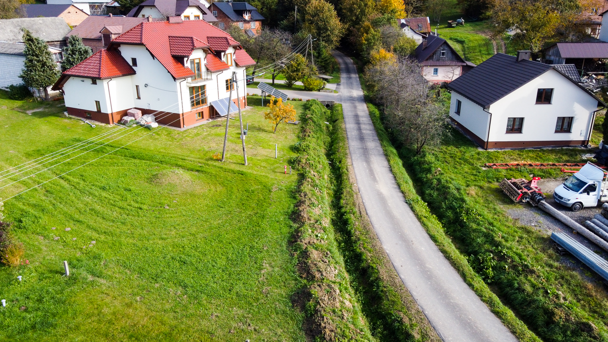 Dwa białe domy z czerwonymi i czarnymi dachami obok asfaltowej drogi, zielona trawa, drzewa i słupki energetyczne z przewodami.