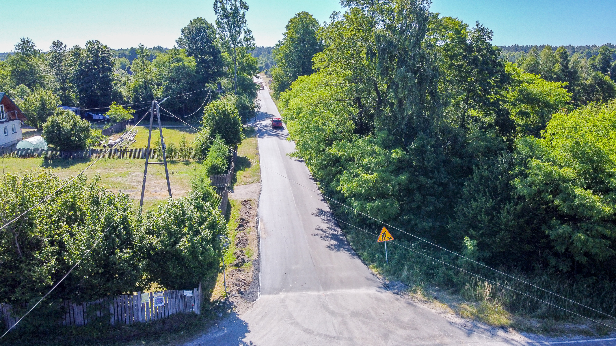 Wąska asfaltowa droga przez wiejską okolicę, po prawej gęste zielone drzewa, po lewej ogrodzone działki z domem, na drodze czerwony samochód i znak ostrzegawczy.