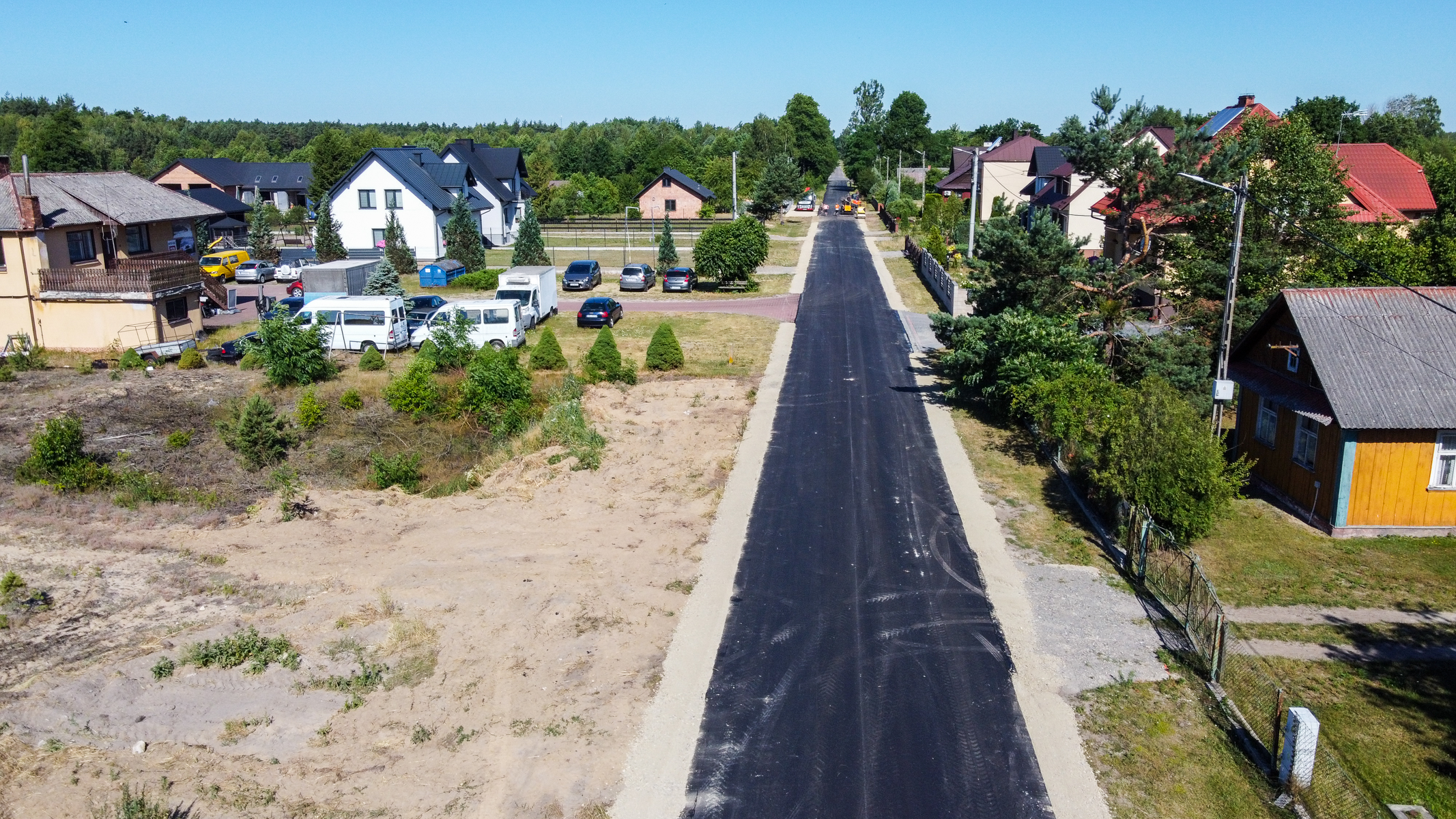 Nowa czarna asfaltowa droga biegnąca przez wiejską zabudowę, po bokach trawniki, krzewy i domy z czerwonymi i szarymi dachami, pod niebieskim niebem.