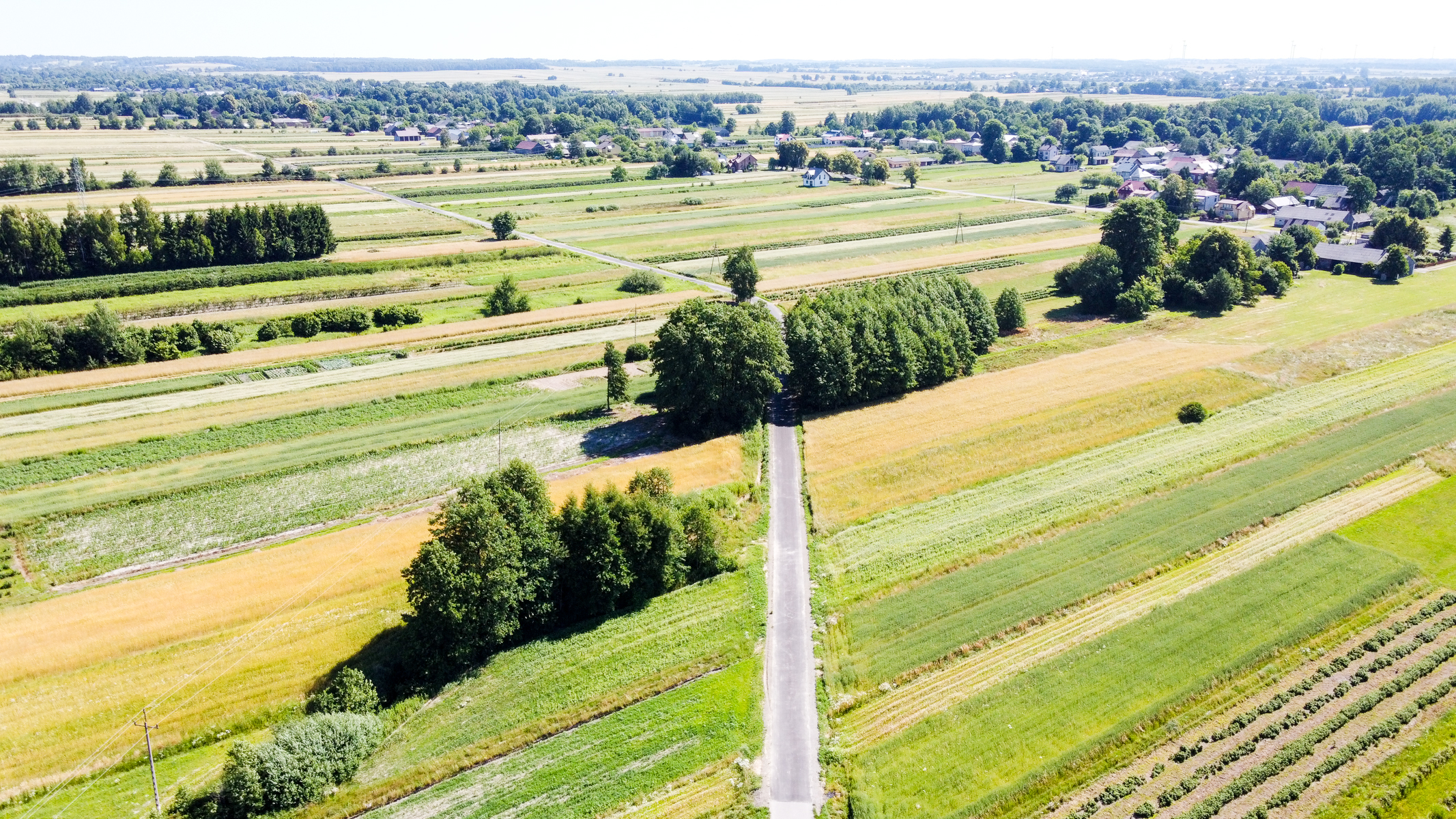 Widok z lotu ptaka na asfaltową drogę przecinającą pola uprawne w różnych odcieniach zieleni i żółci, skupiska drzew oraz zabudowania w tle.