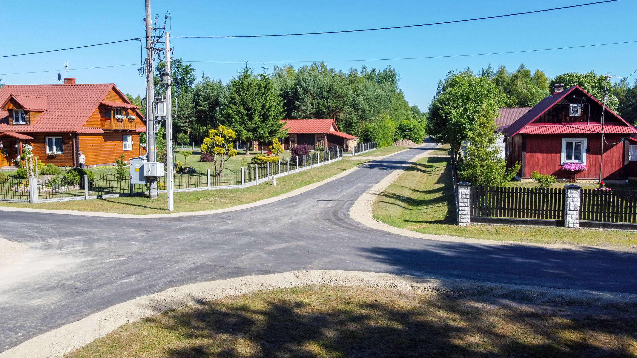 Skrzyżowanie asfaltowych dróg z poboczami z tłucznia, po obu stronach drewniane domy z ogrodami, zieleń drzew i krzewów, niebieskie niebo.