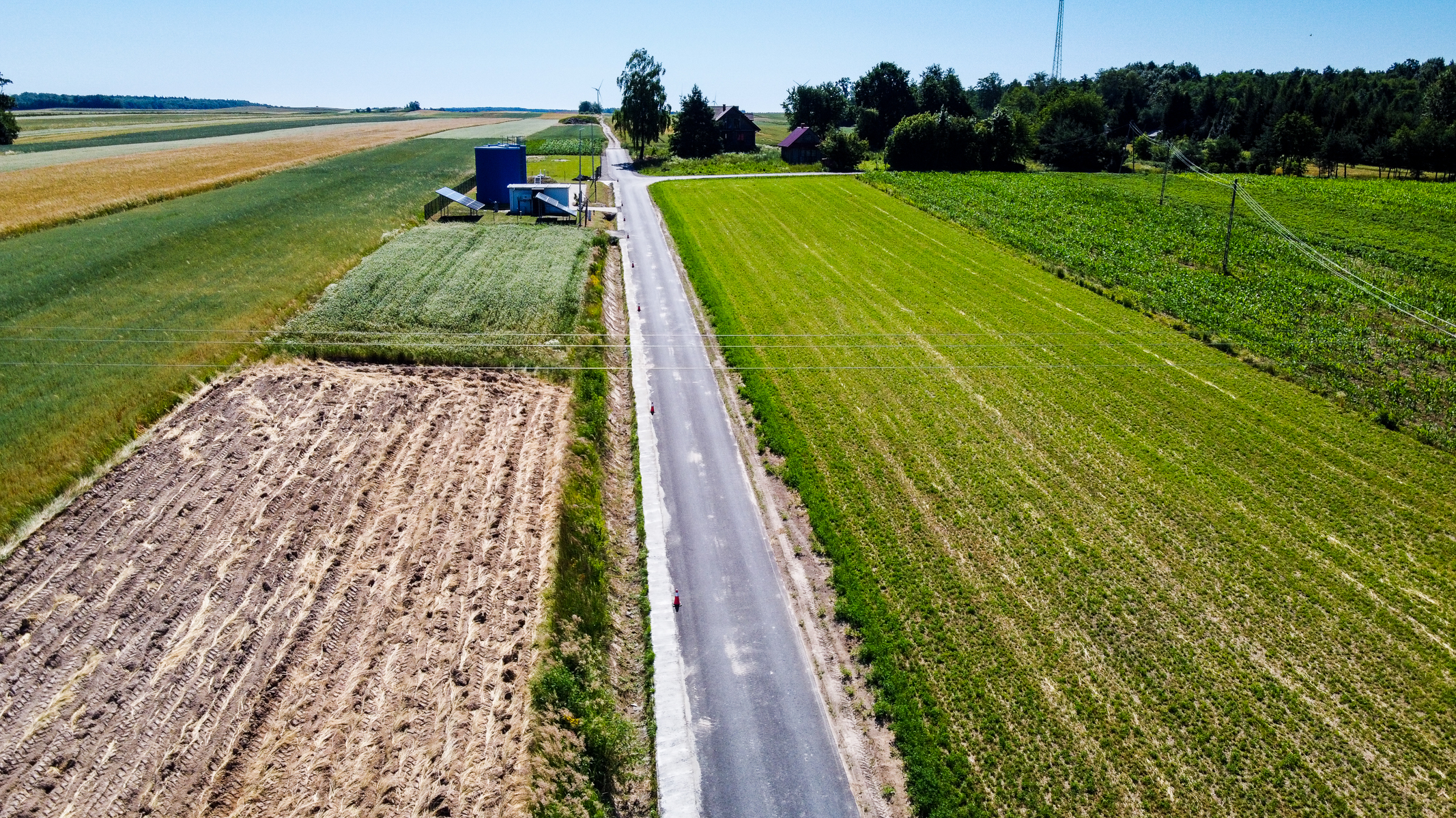 Wąska asfaltowa droga między polami uprawnymi o różnych odcieniach zieleni i brązu, w tle domy, drzewa oraz niebieski zbiornik i panele słoneczne.