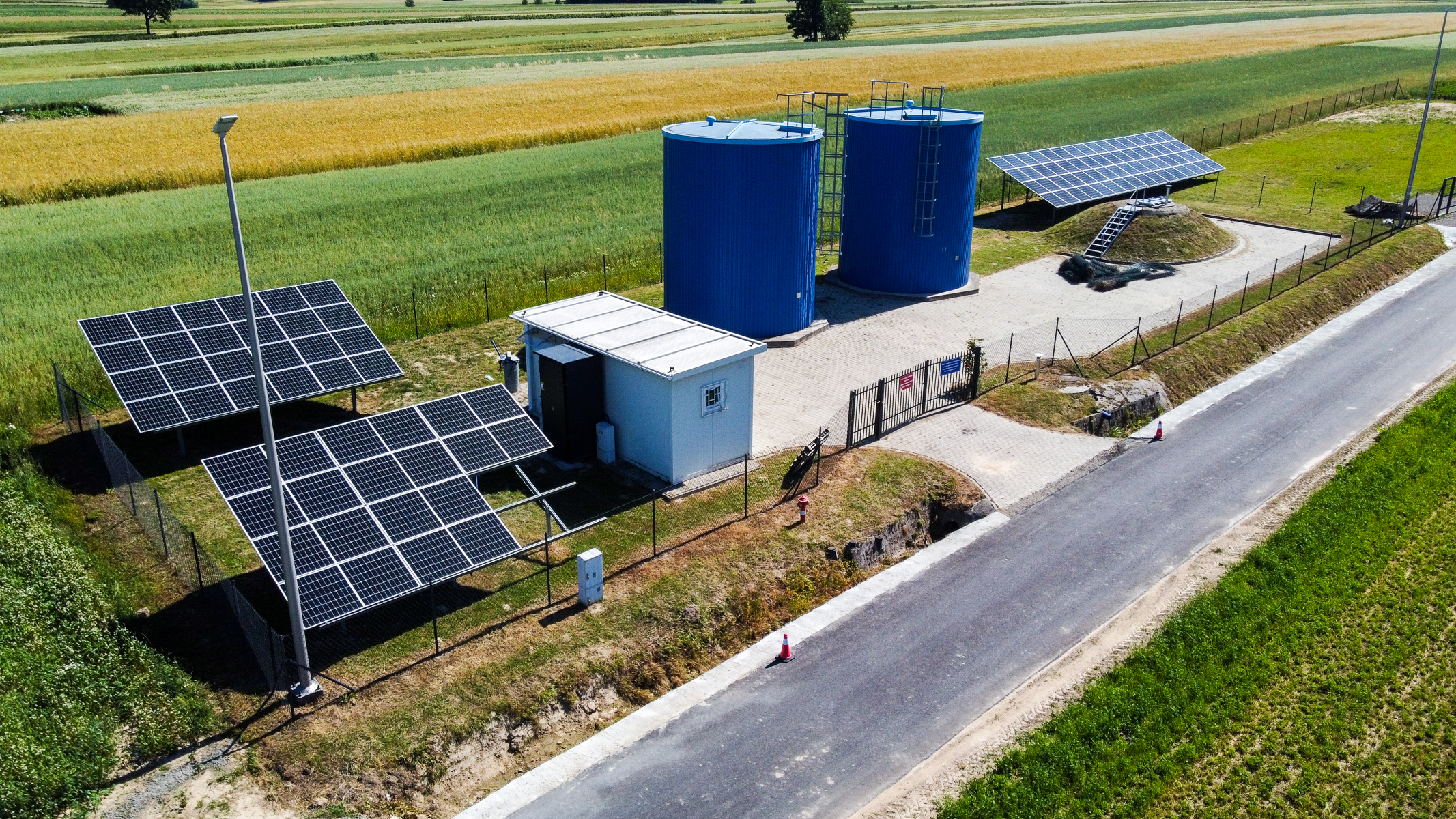 Dwa duże niebieskie zbiorniki, biała budka, panele słoneczne na metalowych stelażach, asfaltowa droga, zielone i żółte pola uprawne w tle.