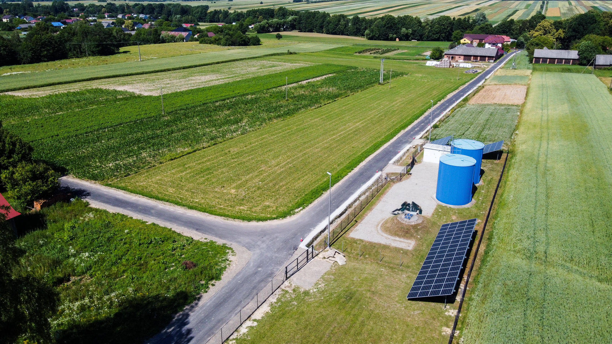 Pole uprawne z zieloną i żółtą trawą, droga asfaltowa, dwa niebieskie zbiorniki, panele słoneczne oraz zabudowania i drzewa w tle.