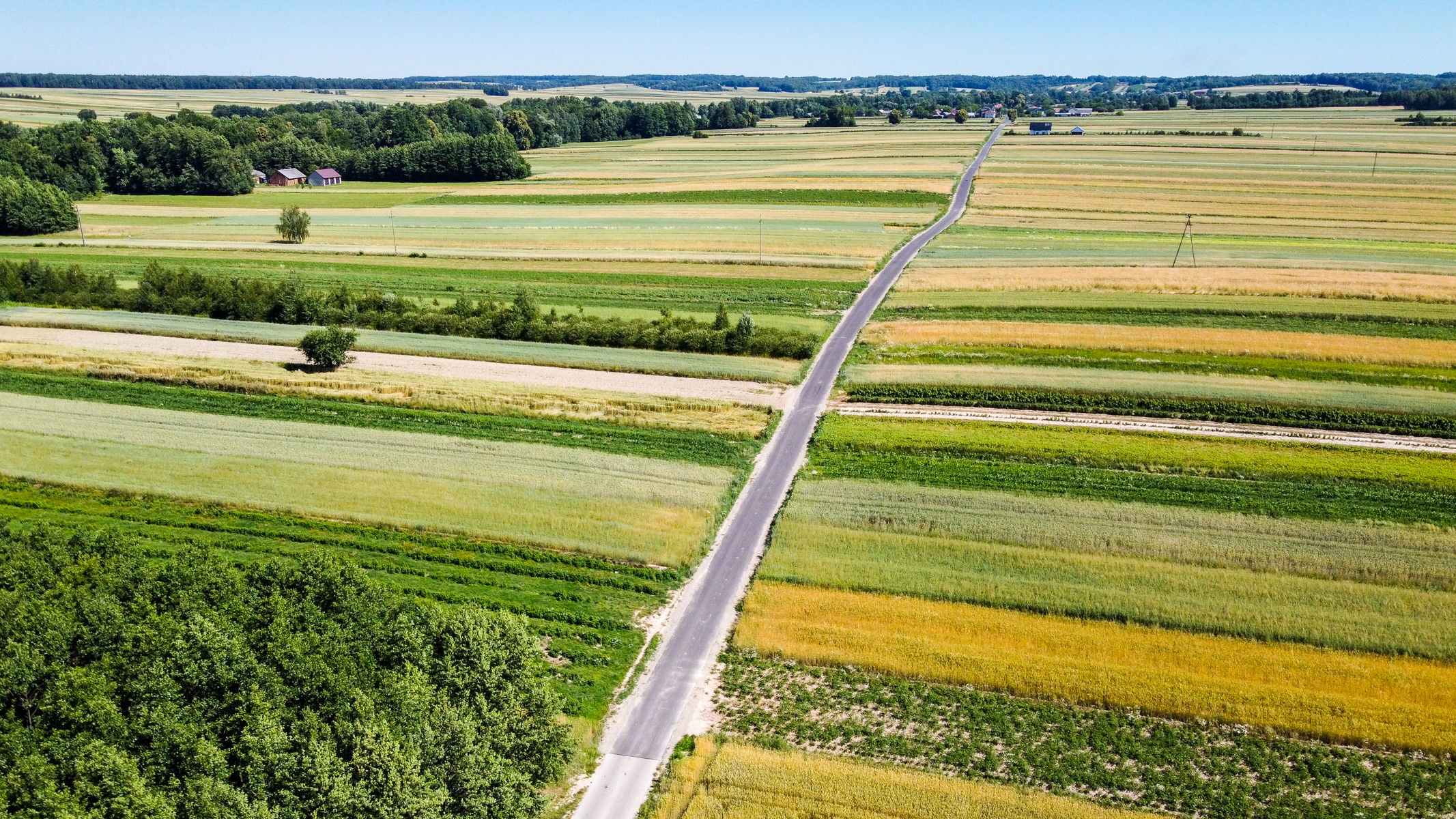 Długa asfaltowa droga przecinająca pola o różnorodnych odcieniach zieleni i żółci, na horyzoncie zalesione wzgórza i kilka budynków pod niebieskim niebem.