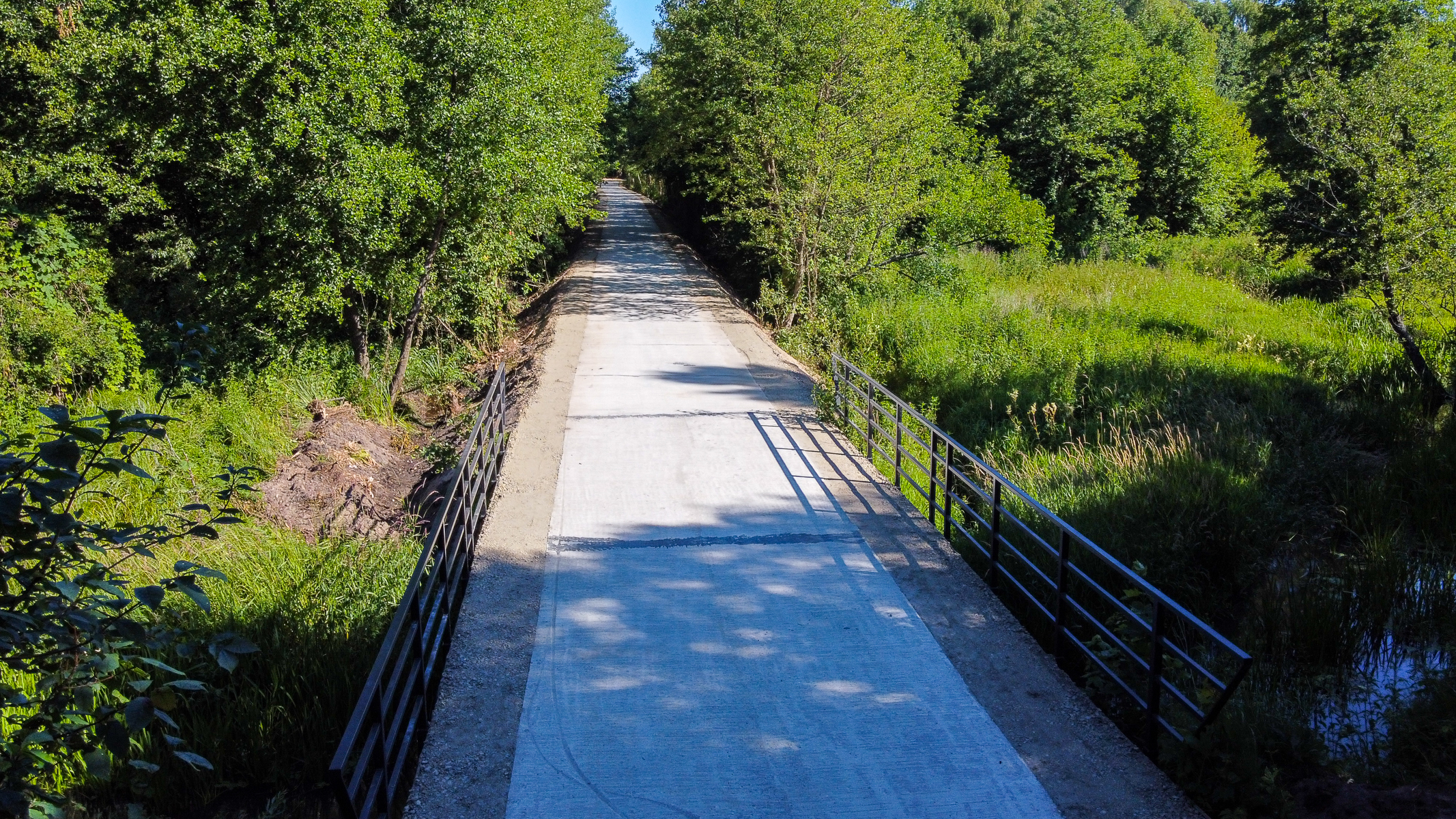 Wąska betonowa droga z metalowymi barierkami po bokach, otoczona gęstą zieloną roślinnością i drzewami, pod błękitnym niebem.