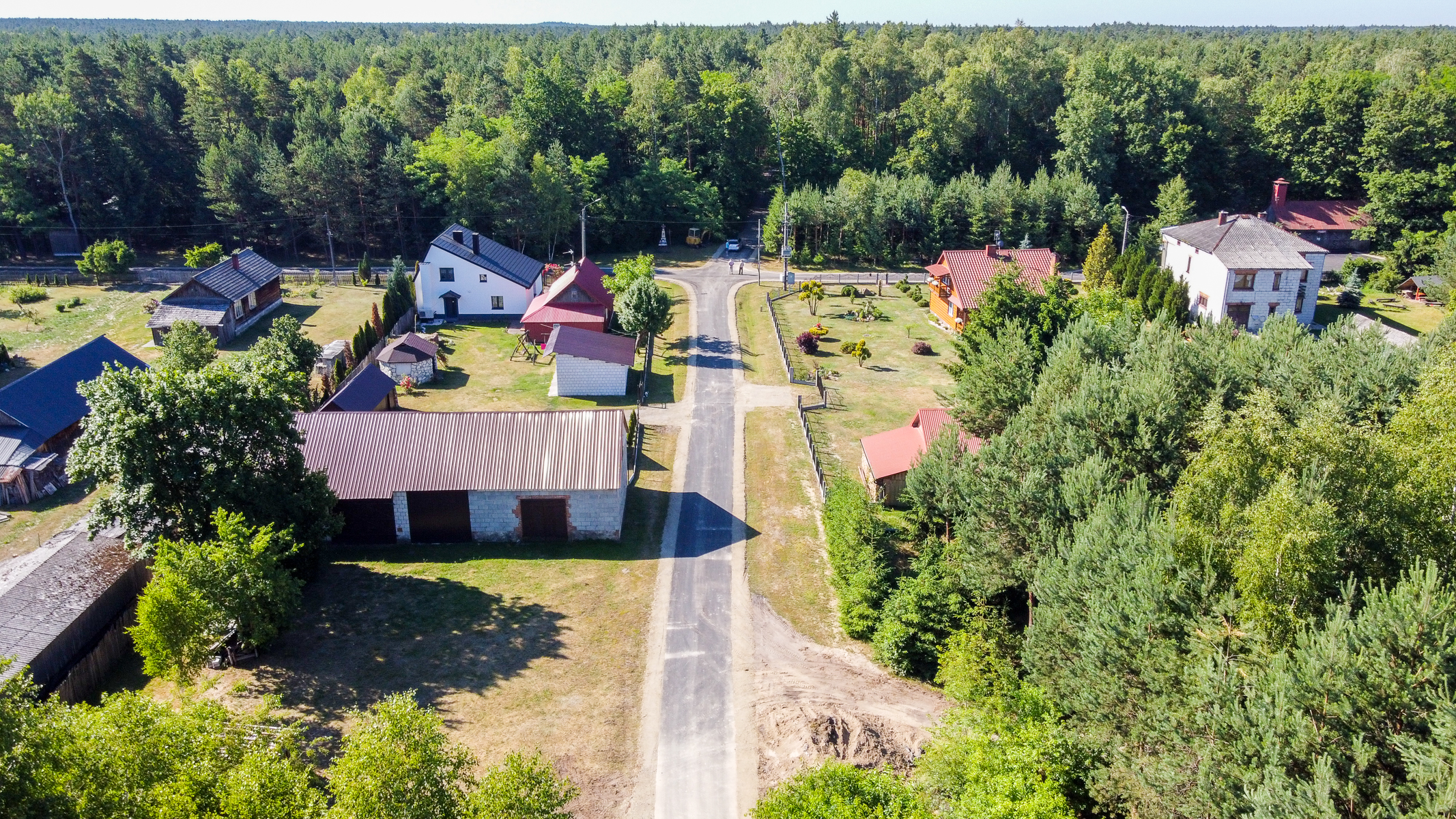 Widok z lotu ptaka na wiejską drogę z domami po obu stronach, otoczoną zielonymi drzewami i lasem w tle, pod jasnym niebem.