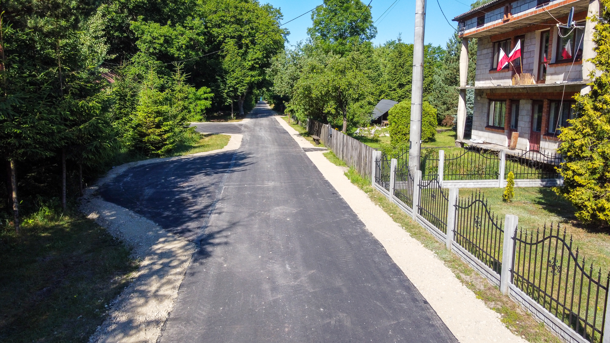 Droga asfaltowa z poboczami z jasnego tłucznia, otoczona zielonymi drzewami, po prawej stronie ogrodzona posesja z budynkiem i dwoma flagami na balkonie.