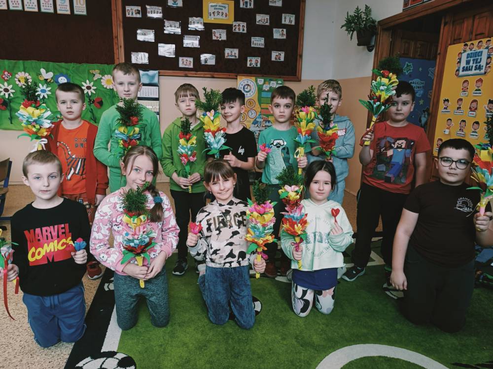 Grupa dzieci w kolorowych ubraniach trzyma wielobarwne palmy wielkanocne, stoją i klęczą na zielonym dywanie w klasie z dekoracjami na ścianach.