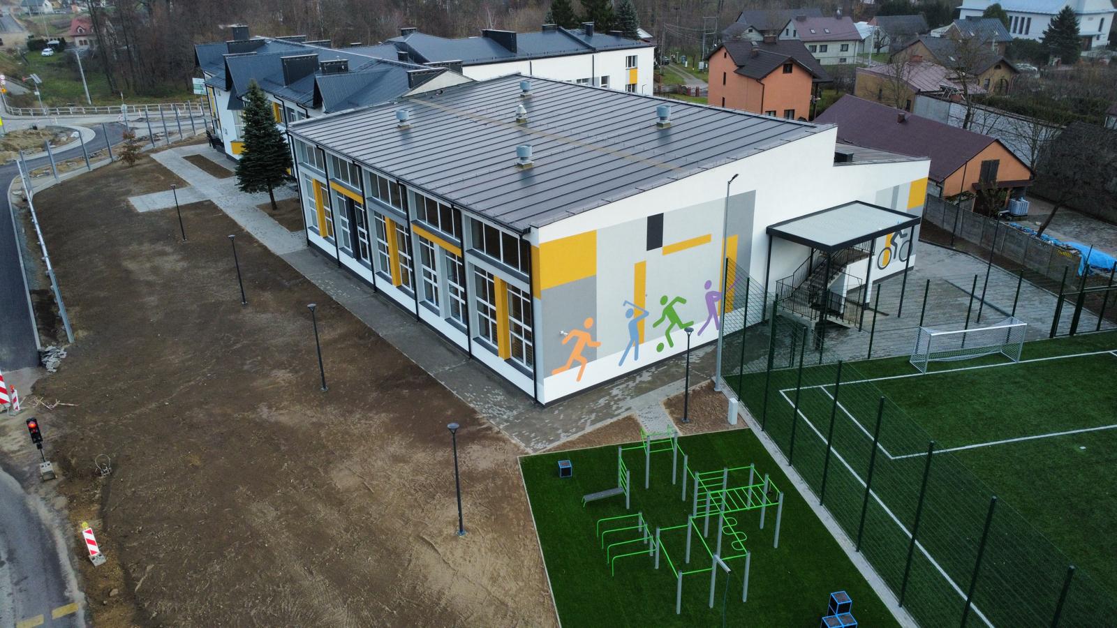 Budynek z szarym dachem i biało-szaro-żółtymi ścianami, kolorowe sylwetki sportowców na ścianie, zielone boisko piłkarskie z białymi liniami, plac do ćwiczeń z zielonym sprzętem.