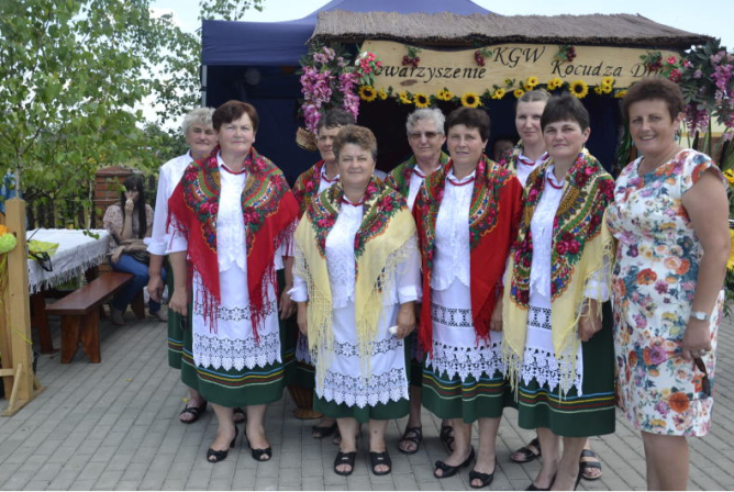 Grupa kobiet w tradycyjnych strojach ludowych z zielonymi spódnicami, białymi bluzkami i kolorowymi chustami, stojąca przed dekorowanym stoiskem z napisem i kwiatami.