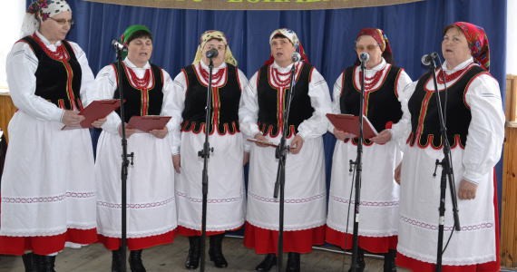 Sześć kobiet w tradycyjnych białych sukniach z czerwonymi spódnicami, czarnych kamizelkach i kolorowych chustach na głowach, stojących przed mikrofonami na tle granatowej kurtyny.