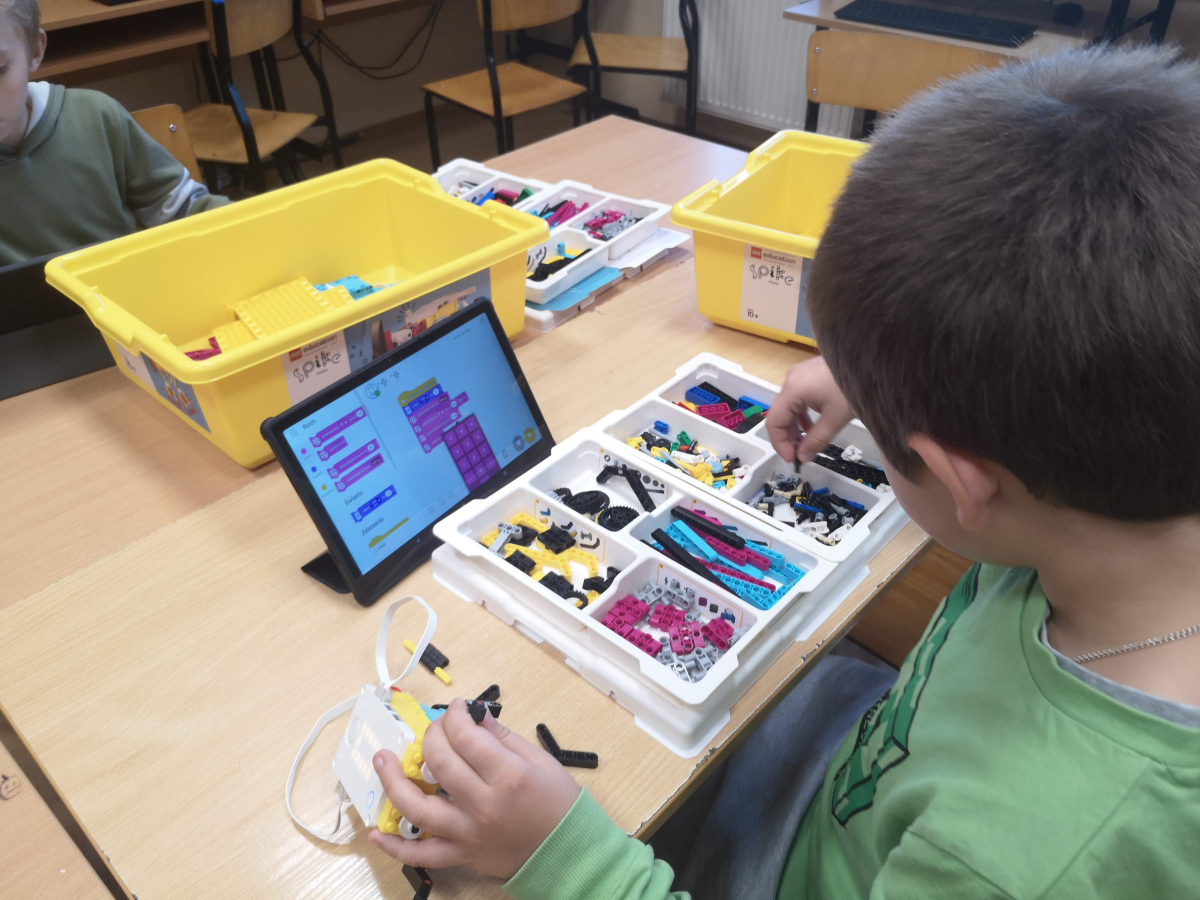 Chłopiec w zielonej bluzie składa elementy konstrukcyjne z klocków LEGO w pojemniku na stole, obok stoi tablet z widocznym programem do kodowania.