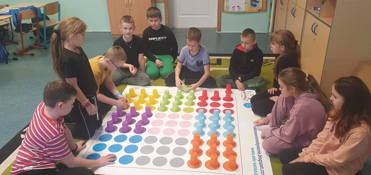 Grupa dzieci siedzi i klęczy wokół dużej planszy z kolorowymi kółkami i plastikowymi kubeczkami w rzędach, w klasie z meblami i tablicą na ścianie.