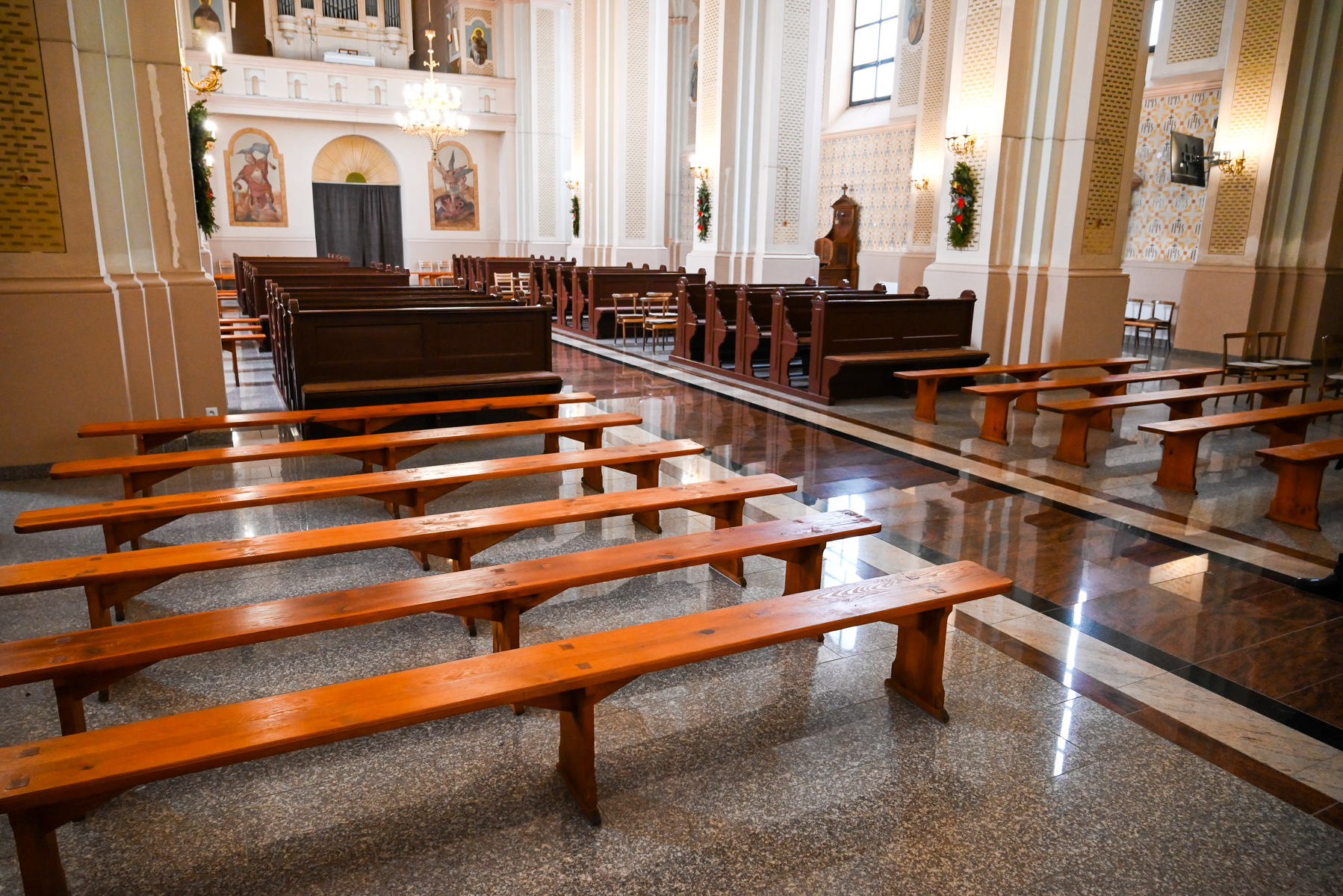 Drewniane ławki i ciemne ławki z oparciami w jasnym wnętrzu kościoła z wysokimi oknami, obrazami i zdobionymi filarami, odbicia światła na marmurowej posadzce.