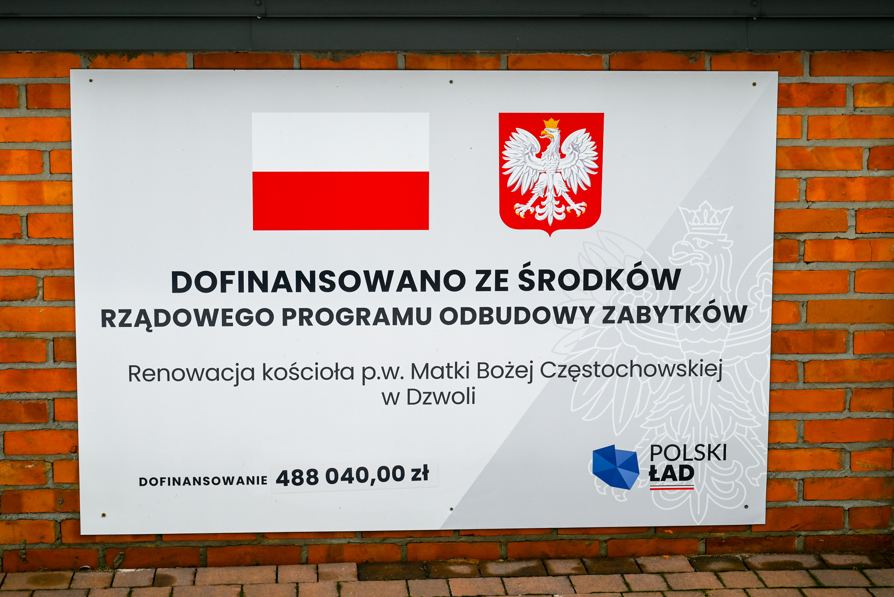Tablica informacyjna na tle ceglanej ściany, biały prostokąt z flagą Polski, herbem orła i tekstem o dofinansowaniu renowacji kościoła w Dzwoli kwotą 488 040 zł.