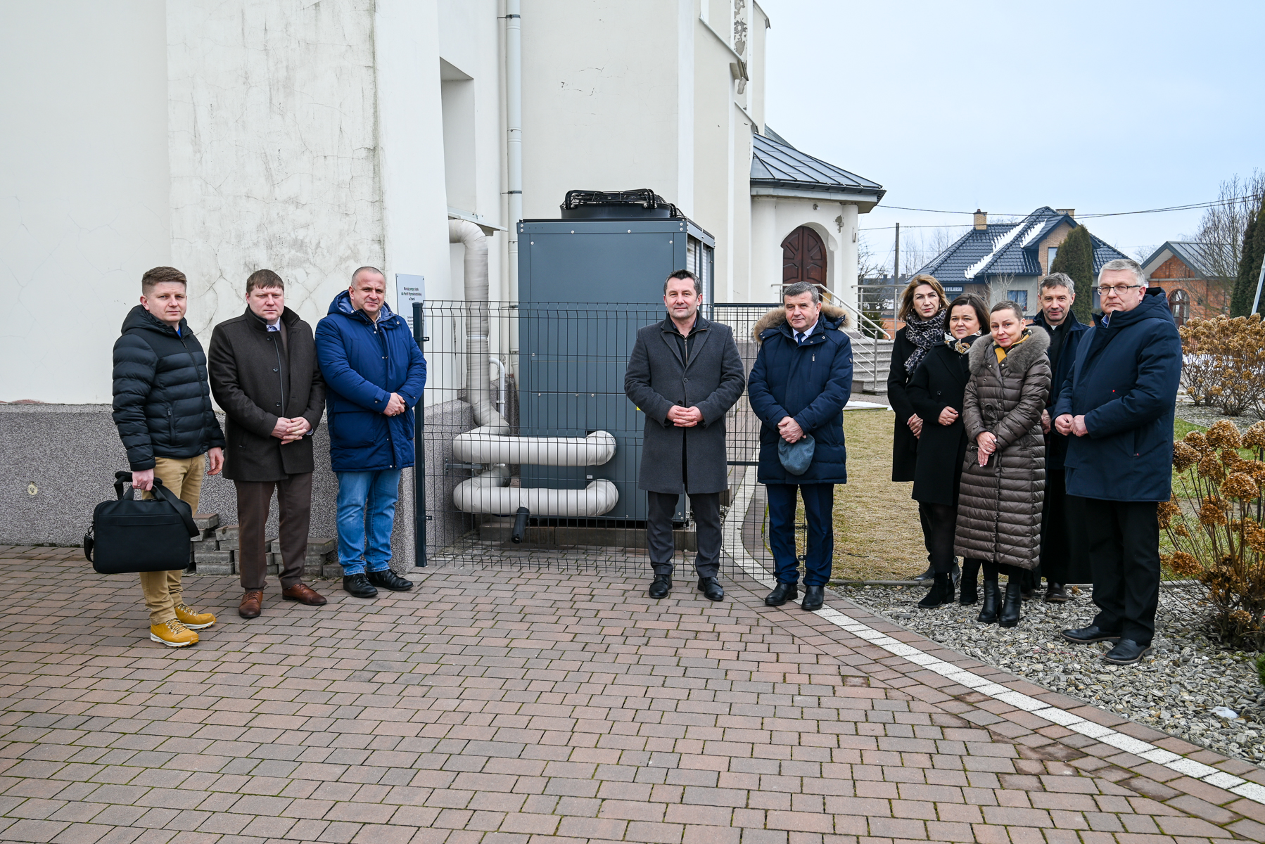 Dziesięć osób w zimowych ubraniach stoi na kostce brukowej i kamiennej nawierzchni przed budynkiem z białą elewacją i metalową instalacją z rurami.