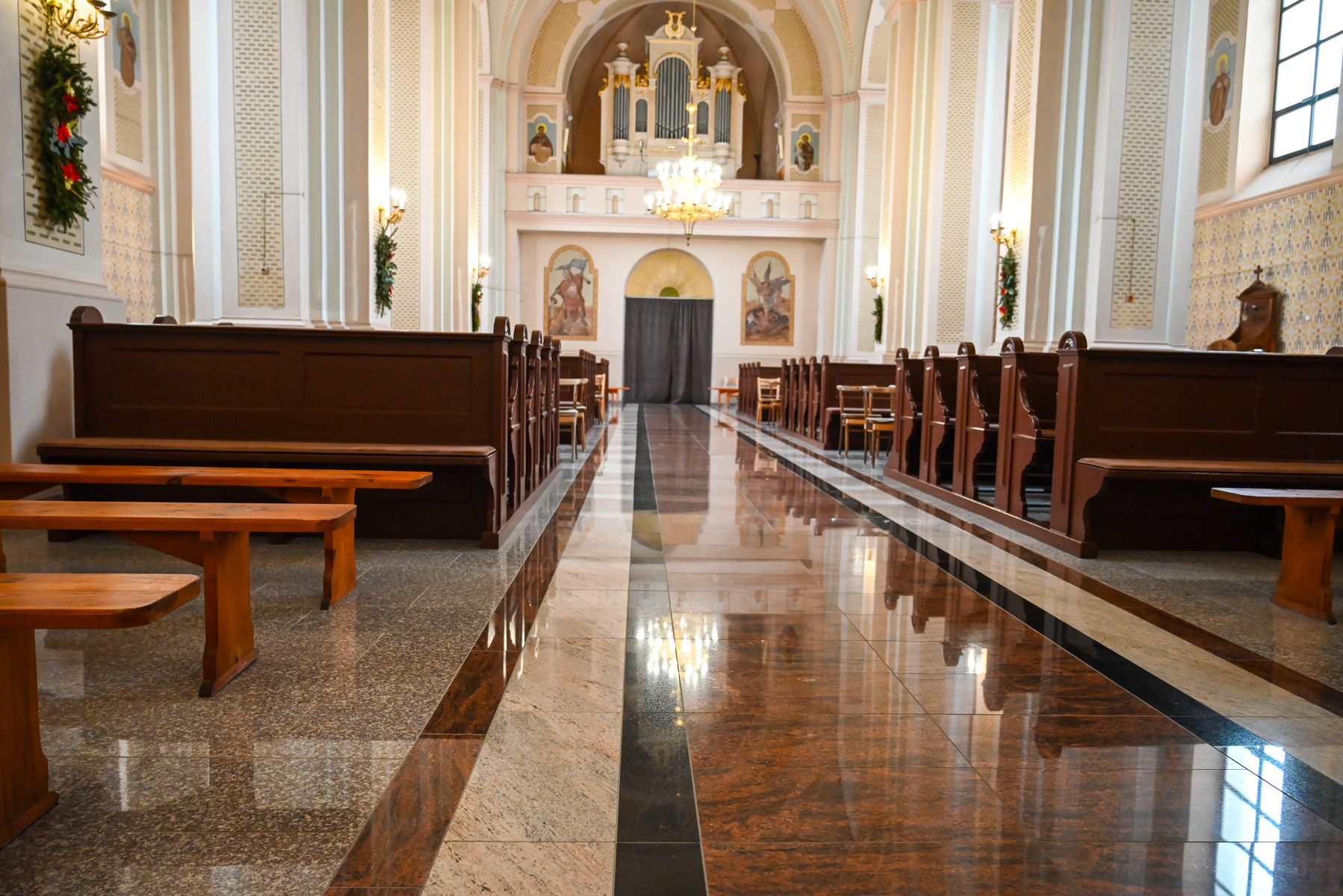 Wnętrze jasnej świątyni z ciemnymi drewnianymi ławkami po bokach i błyszczącą, marmurową posadzką w odcieniach brązu i beżu, na końcu organ i żyrandol.