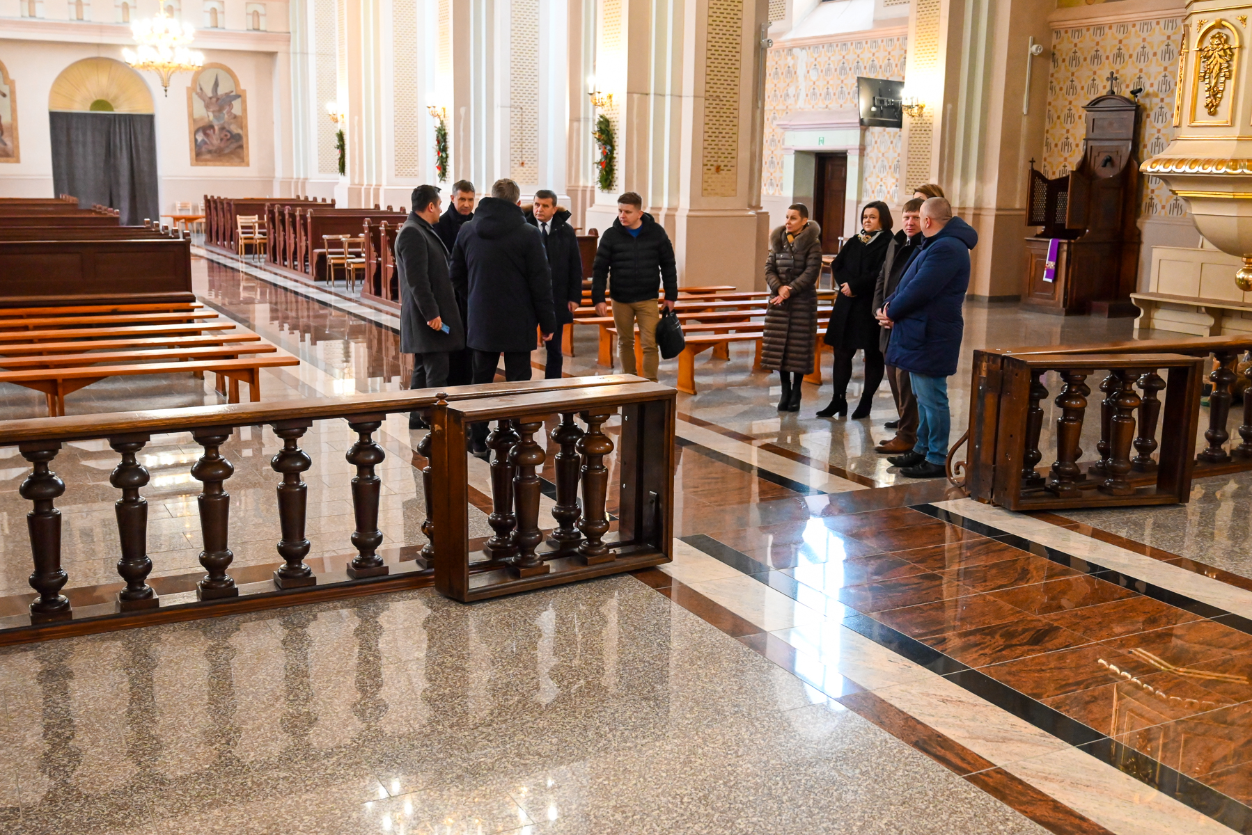 Grupa ośmiu osób w ciemnych płaszczach stoi na marmurowej podłodze w jasnym wnętrzu z drewnianymi ławkami i ozdobionymi kolumnami.