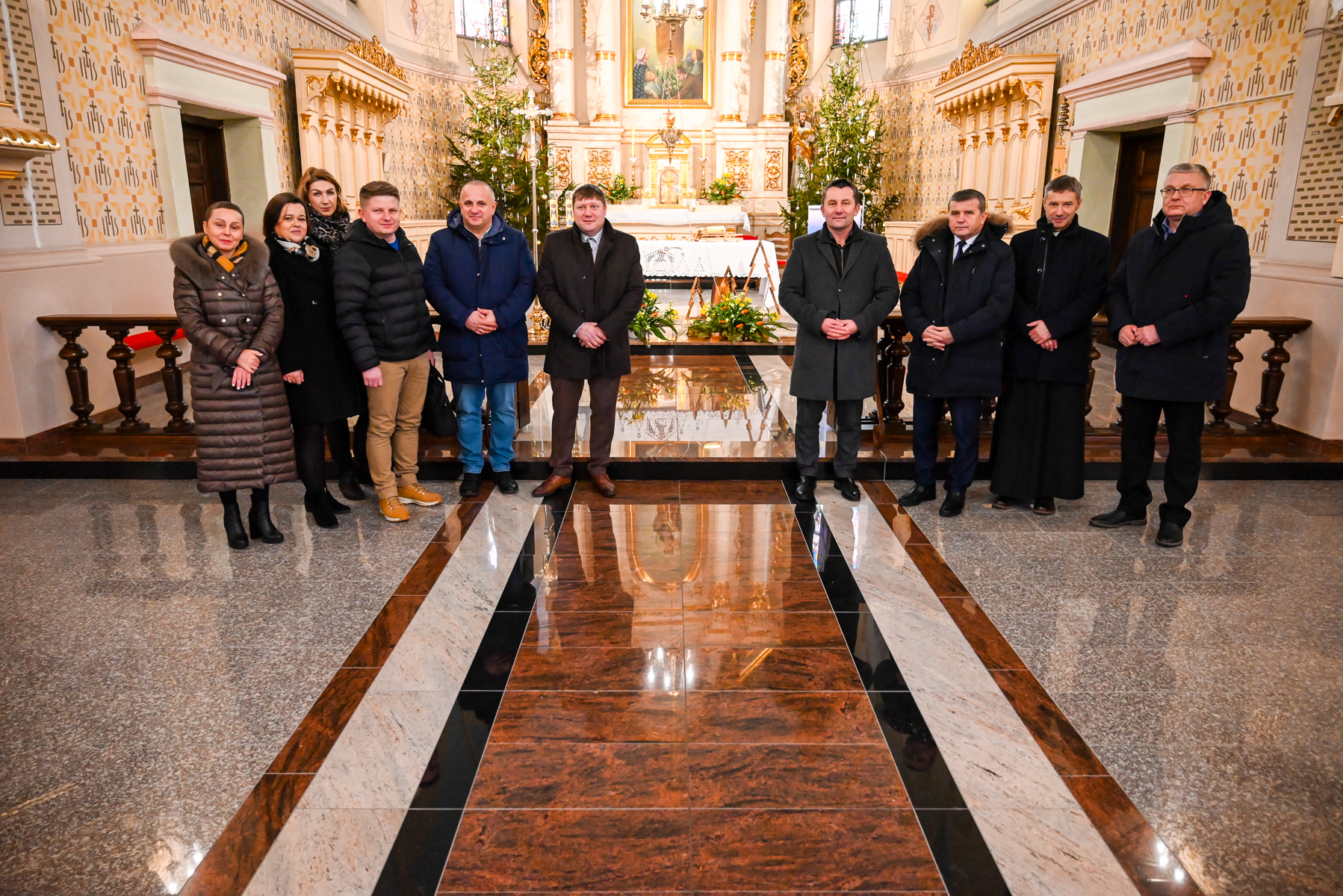 Dziesięć osób w zimowych płaszczach stoi na marmurowej posadzce w jasnym, zdobionym wnętrzu kościoła z choinkami i ołtarzem w tle.