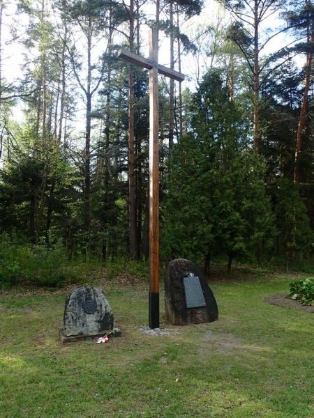 Drewniany krzyż na trawiastym placu, obok dwa kamienie z tablicami pamiątkowymi, w tle drzewa iglaste i liściaste.