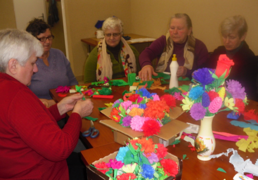 Cztery starsze kobiety siedzą przy stole z kolorowymi papierowymi kwiatami i materiałami do rękodzieła w ciepłym, beżowym pomieszczeniu.
