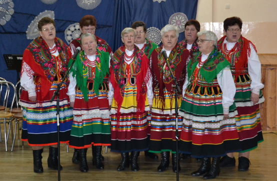 Grupa ośmiu kobiet w kolorowych ludowych strojach z haftowanymi chustami, spódnicami i koralami, stojąca na drewnianej podłodze przed niebieską zasłoną.