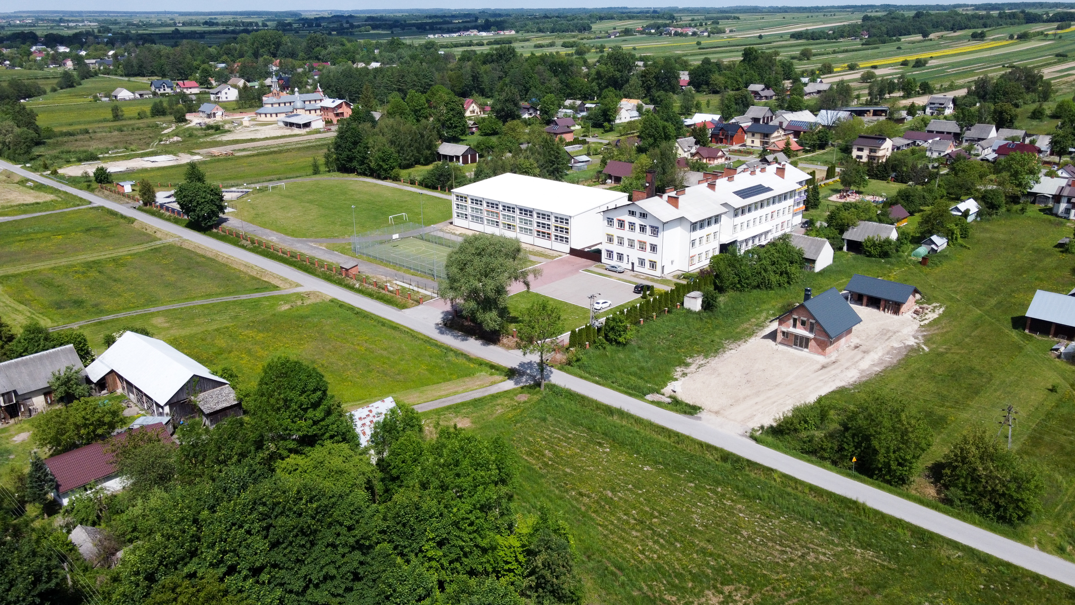 Widok z lotu ptaka na wiejską zabudowę z białym budynkiem szkoły, boiskiem sportowym, drogami, zielonymi polami i drzewami.