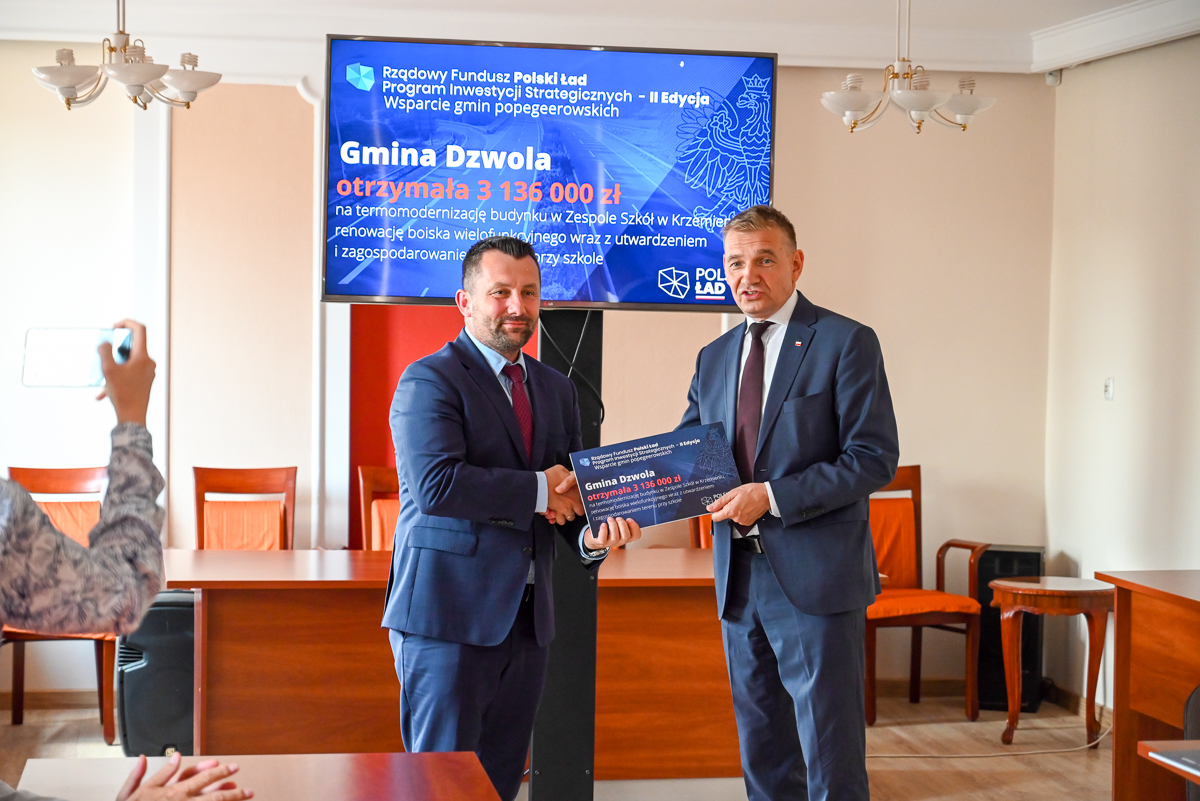 Dwóch mężczyzn w garniturach ściska dłonie, trzymając tablicę, za nimi ekran z niebieskim tłem i tekstem, w tle drewniane krzesła i stoły, po lewej osoba robiąca zdjęcie telefonem.