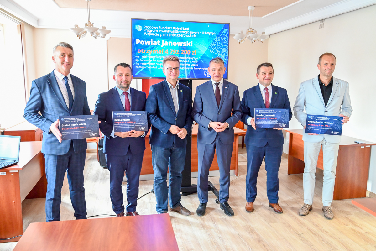 Sześciu mężczyzn w garniturach i marynarkach stoi w jasnym pokoju, trzymając tablice z napisami o dofinansowaniu, za nimi ekran z informacjami w kolorze niebieskim.