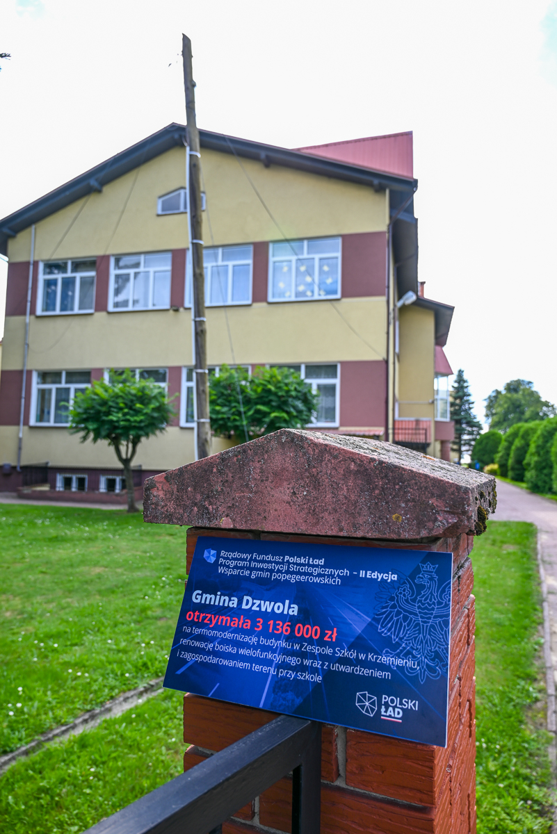 Tablica informacyjna o dofinansowaniu w języku polskim na ceglanym słupku z czerwonym daszkiem, w tle budynek z żółto-brązowymi ścianami i zielony trawnik.