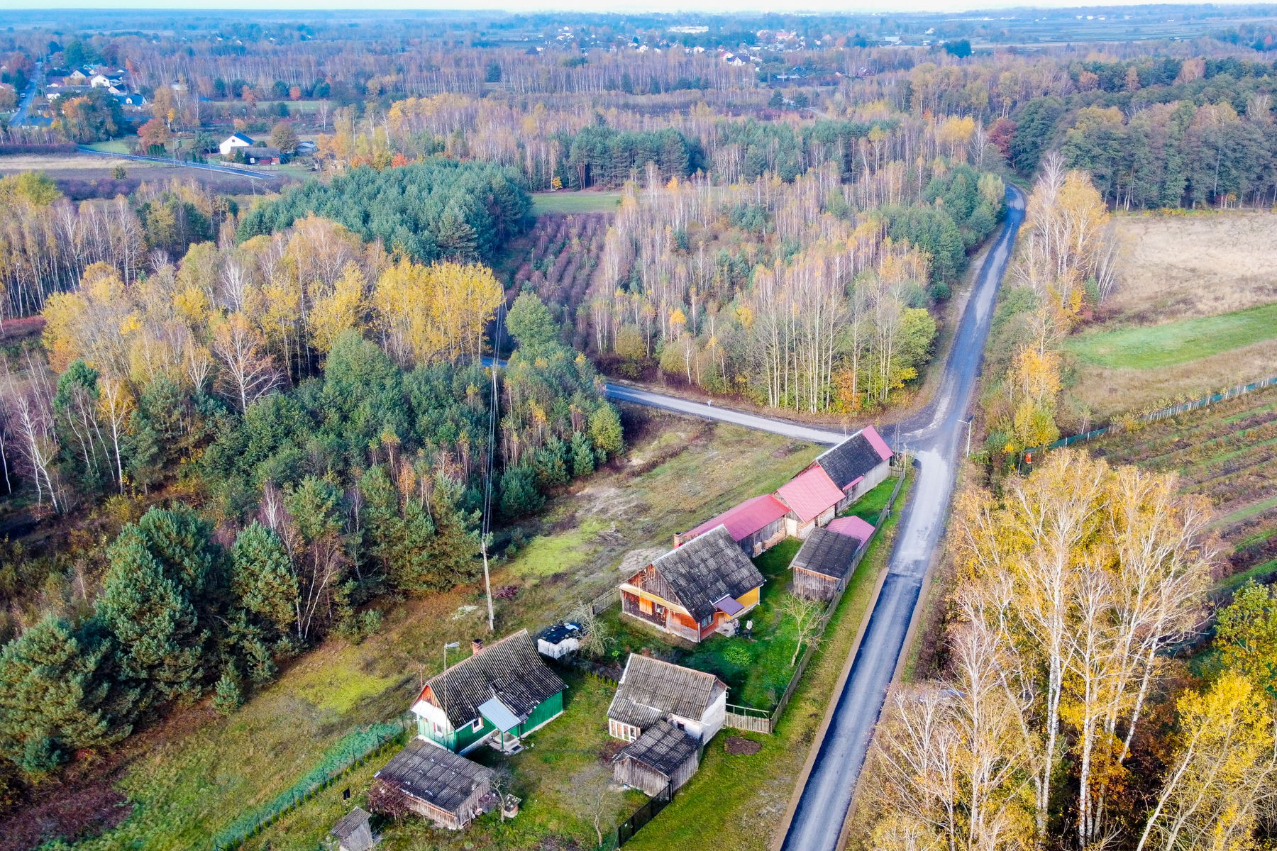 Widok z lotu ptaka na wiejskie domy z dachami w odcieniach szarości i czerwieni, otoczone jesiennym lasem z żółtymi i zielonymi drzewami oraz dwoma drogami asfaltowymi.