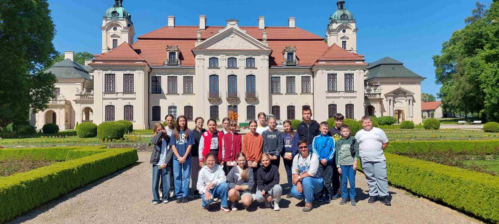 Grupa osób pozująca na tle dużego pałacu z czerwonym dachem, białą fasadą i dwoma wieżami, otoczona zielonymi żywopłotami i drzewami pod niebieskim niebem.