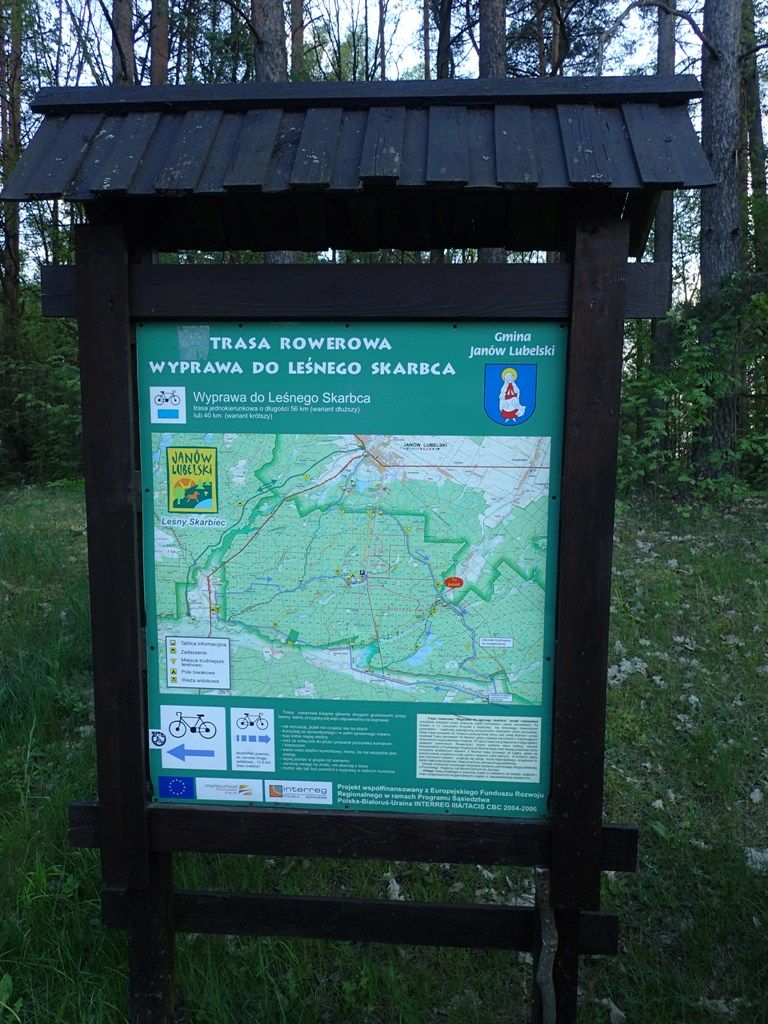 Mapa trasy rowerowej z nazwą "Wyprawa do Leśnego Skarbca", zielone tło, czarny drewniany stojak na tle lasu i trawy.