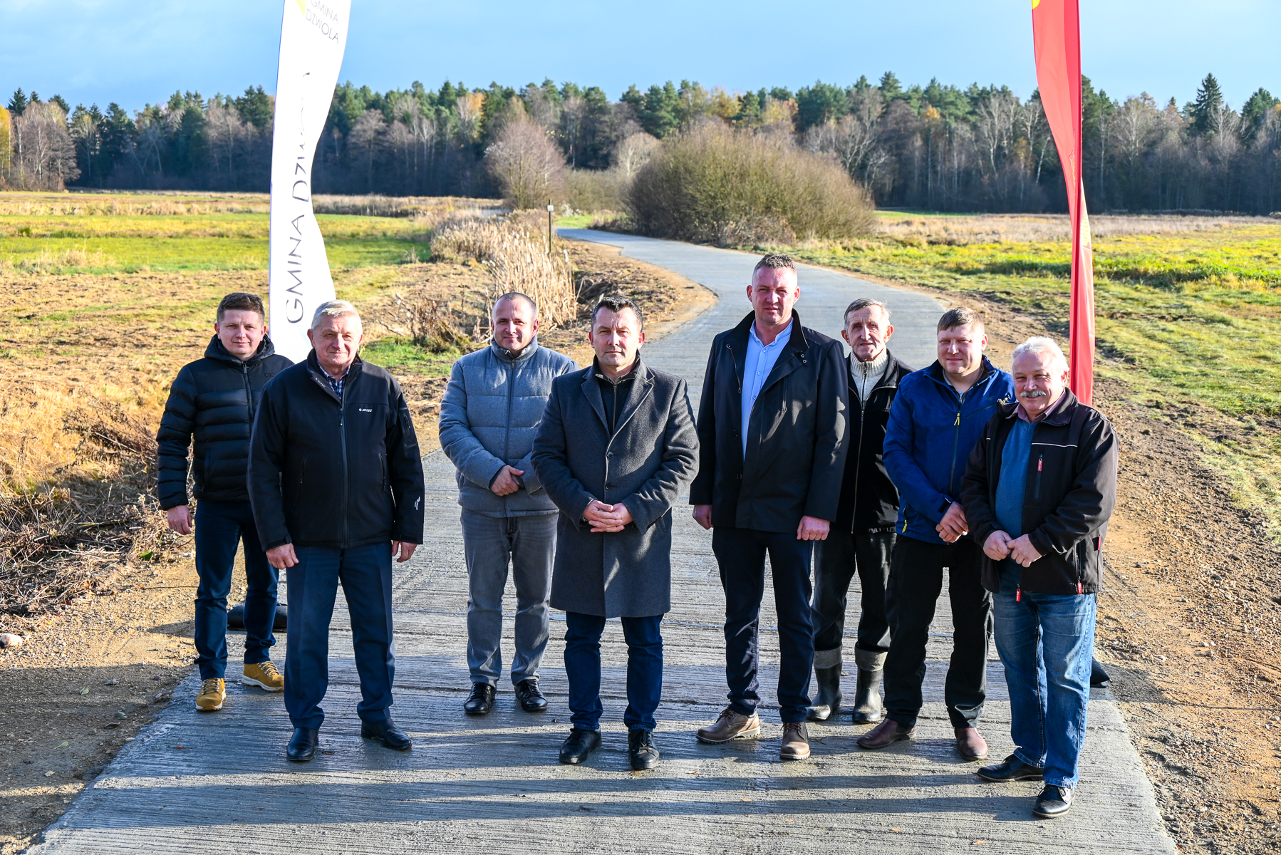 Grupa ośmiu mężczyzn w jesiennym krajobrazie, stojących na betonowej drodze, w tle pola i las, po bokach dwie pionowe flagi.