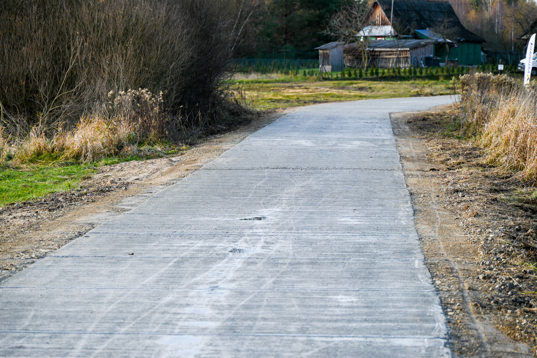 Droga betonowa wśród suchych traw i krzewów, w tle drewniany dom z ciemnym dachem, poranne światło, lekko zagięta ścieżka.