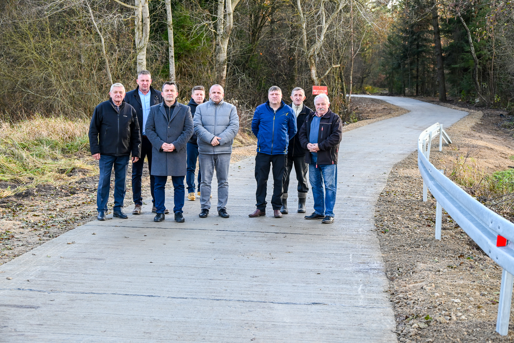 Osiem mężczyzn stojących na betonowej drodze, po prawej stronie metalowa barierka, w tle drzewa i tablica z napisem.