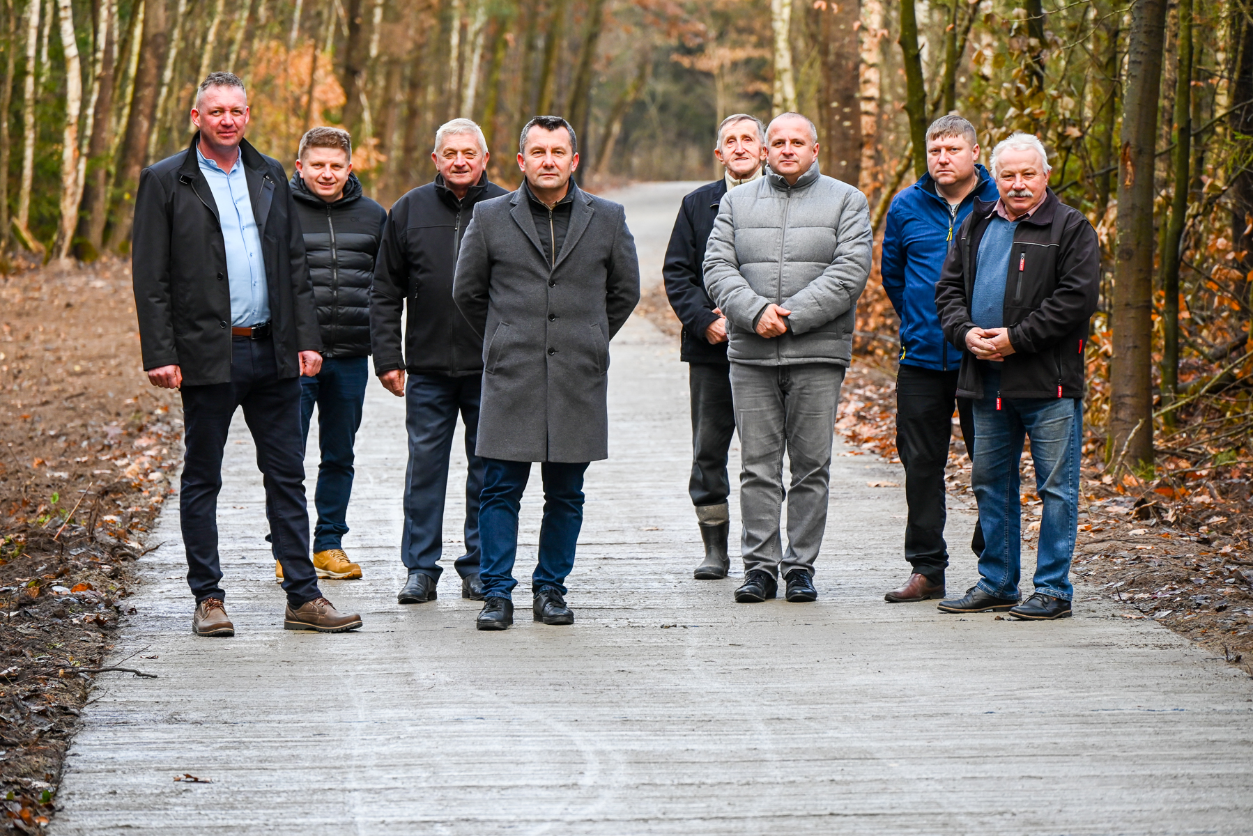 Osiem stojących mężczyzn w jesiennym lesie na betonowej drodze, ubranych w kurtki i płaszcze w ciemnych i szarych kolorach.