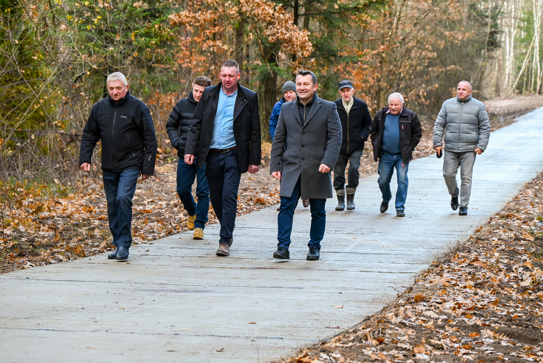 Osiem mężczyzn w jesiennych kurtkach idzie szeroką, szarą ścieżką leśną, wokół suche liście i drzewa z brunatnymi liśćmi.