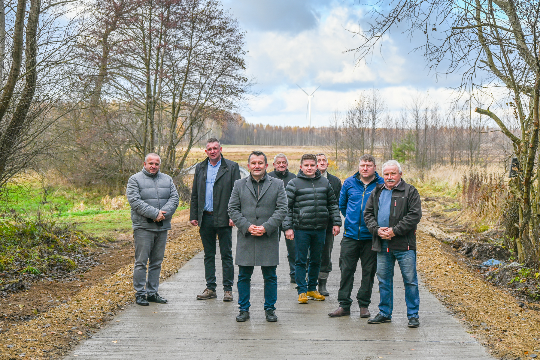 Osiem mężczyzn w kurtkach i płaszczach stoi na nowej drodze otoczonej jesiennymi drzewami i polami pod pochmurnym niebem.