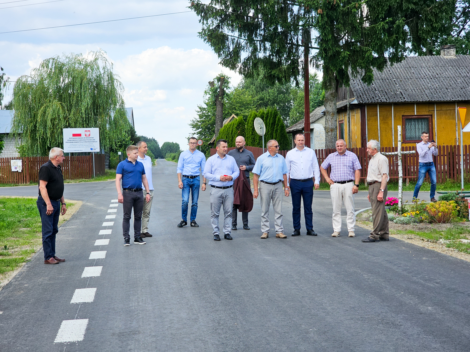 Grupa mężczyzn stoi na świeżo wyasfaltowanej drodze wiejskiej, w tle domy, drzewa i tablice informacyjne. Jeden mężczyzna fotografuje z boku.