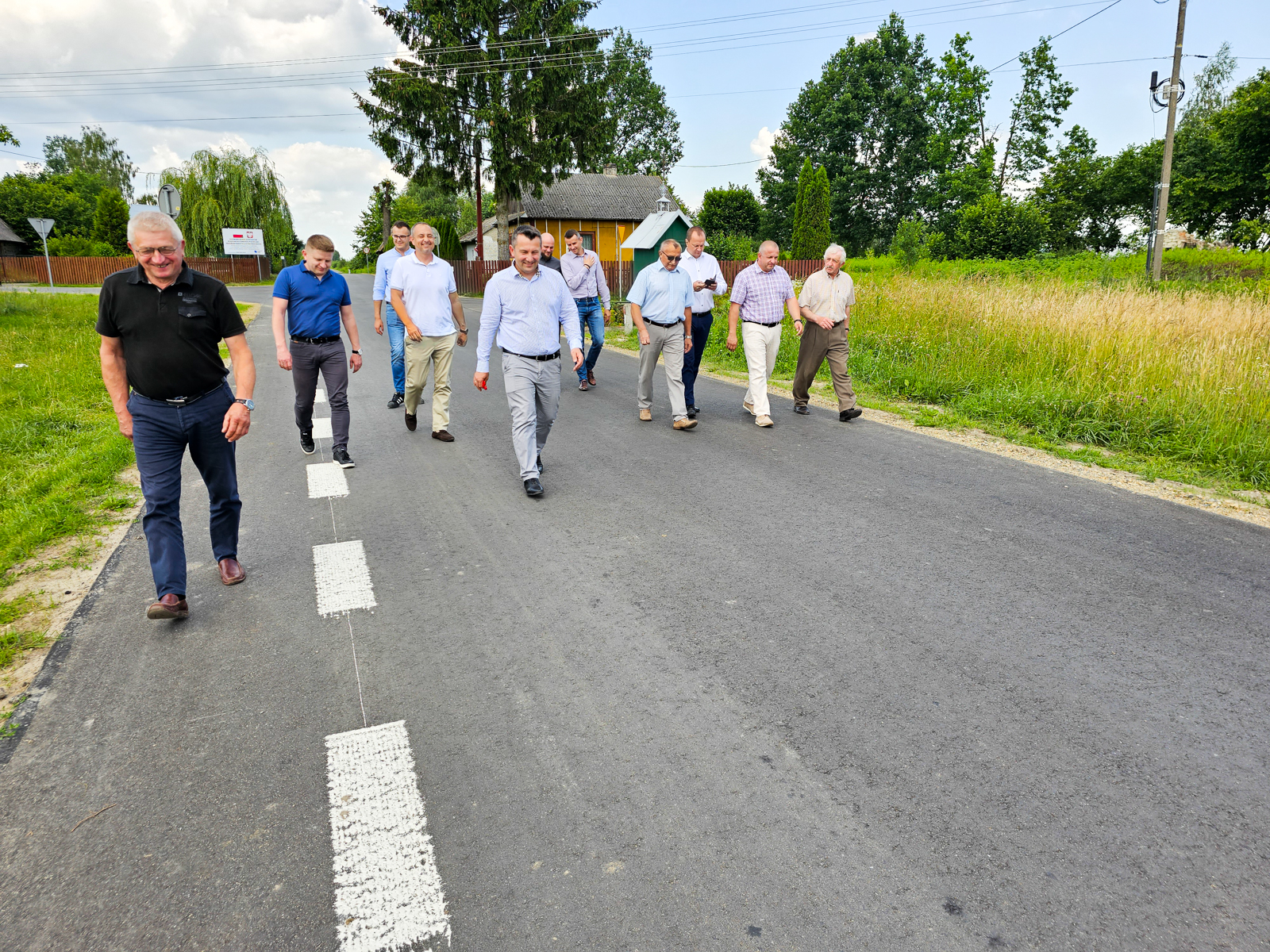 Dziesięciu mężczyzn w letnich ubraniach idzie po asfaltowej drodze, po bokach trawa i drzewa, w tle drewniany płot i dom, niebo z chmurami.