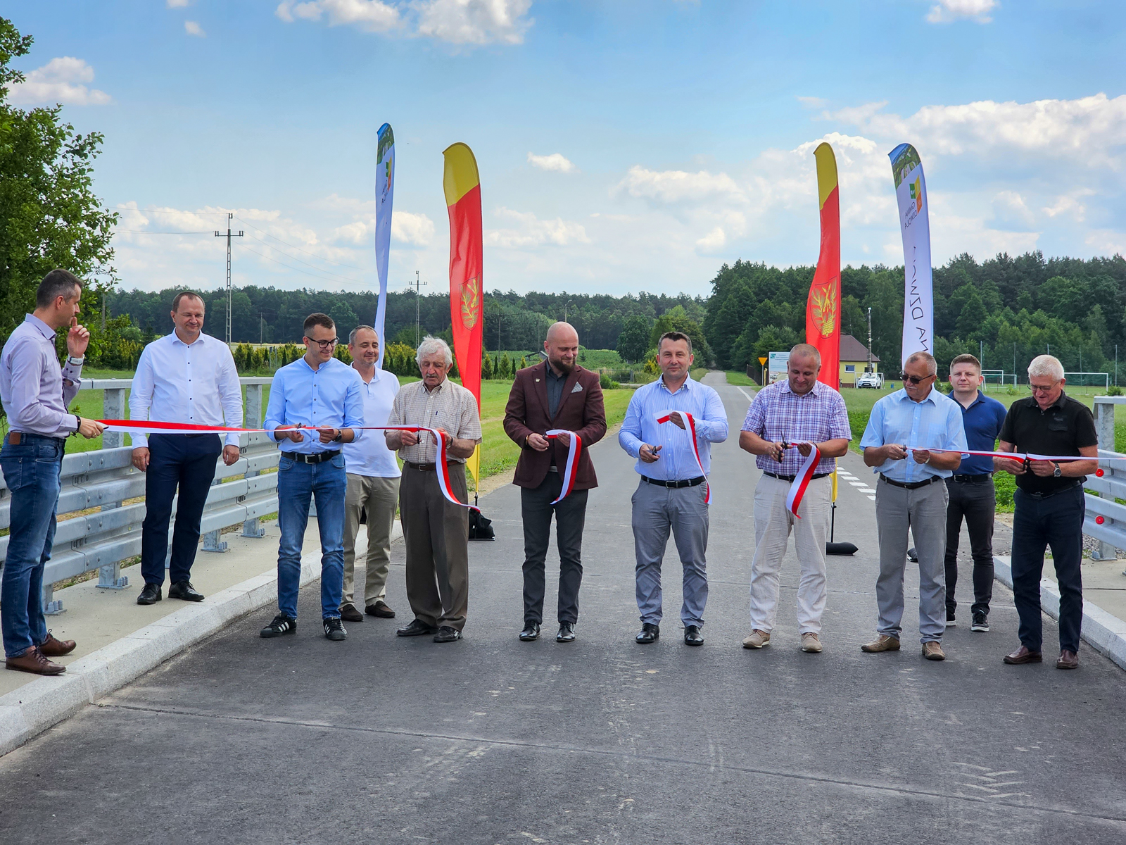 Dziesięć osób w różnorodnych ubraniach stoi na nowej drodze z szarfą w biało-czerwonych barwach, dwie flagi i zieleń w tle pod niebieskim niebem.
